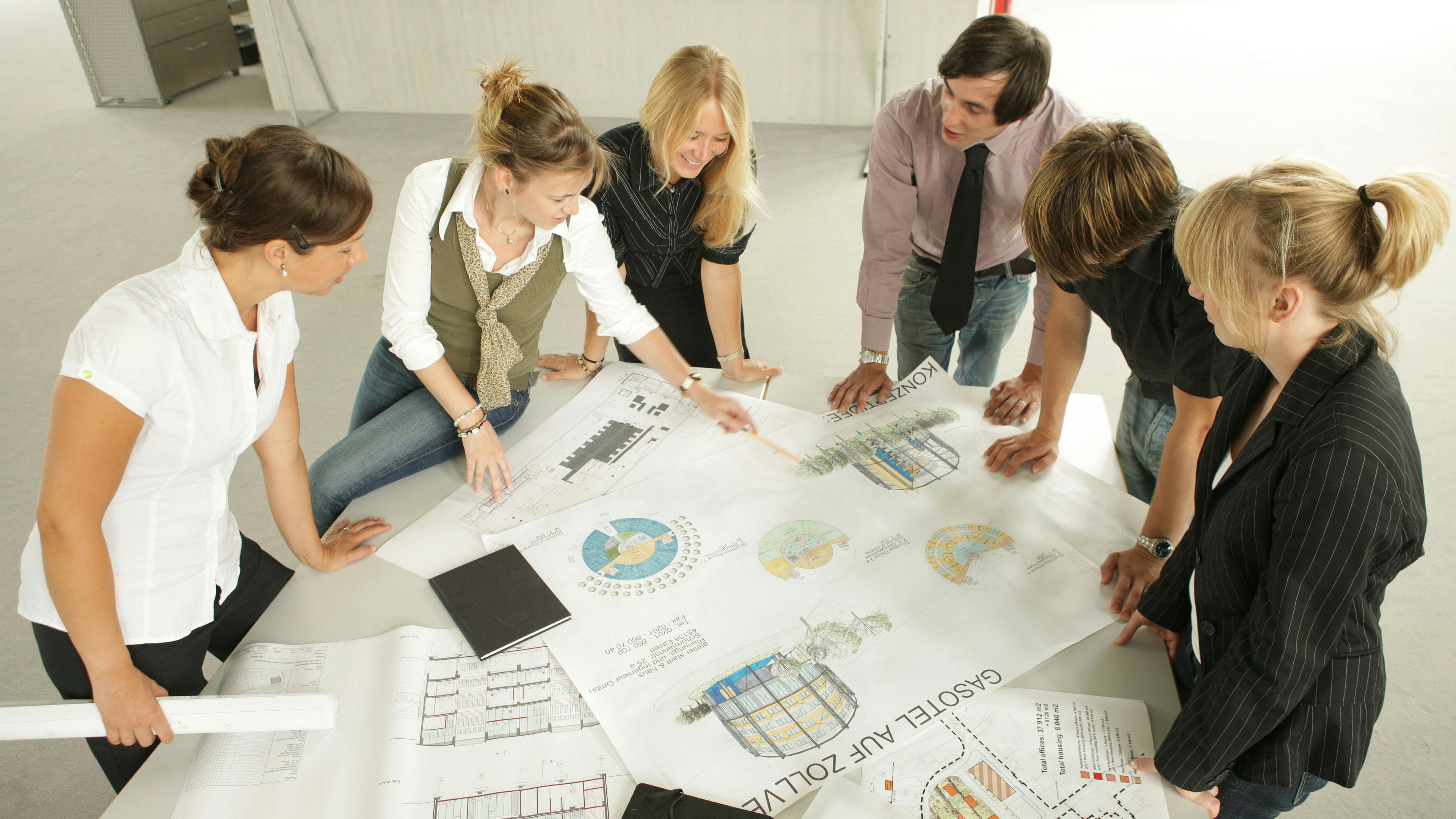 Eine Gruppe von sechs jungen Architekten oder Stadtplanern steht um einen großen Tisch und diskutiert über Entwürfe für ein Bauprojekt auf dem UNESCO-Welterbe Zollverein. Auf dem Tisch liegen mehrere großformatige Architekturpläne, darunter ein farbiger Entwurf mit der Aufschrift Gasotel auf Zollverein sowie schematische Darstellungen und Grundrisse. Eine Frau zeigt mit einem Bleistift auf eine bestimmte Stelle der Zeichnung, während die anderen aufmerksam zuhören und mitdiskutieren. Die Gruppe wirkt engagiert und arbeitet in einem modernen, lichtdurchfluteten Raum mit Betonboden und einer offenen, industriellen Atmosphäre – passend zum industriellen Erbe der Zeche Zollverein. Die Szene vermittelt eine kreative und kollaborative Arbeitsweise im Rahmen eines Projekts zur Umnutzung historischer Industrieanlagen auf dem Gelände.