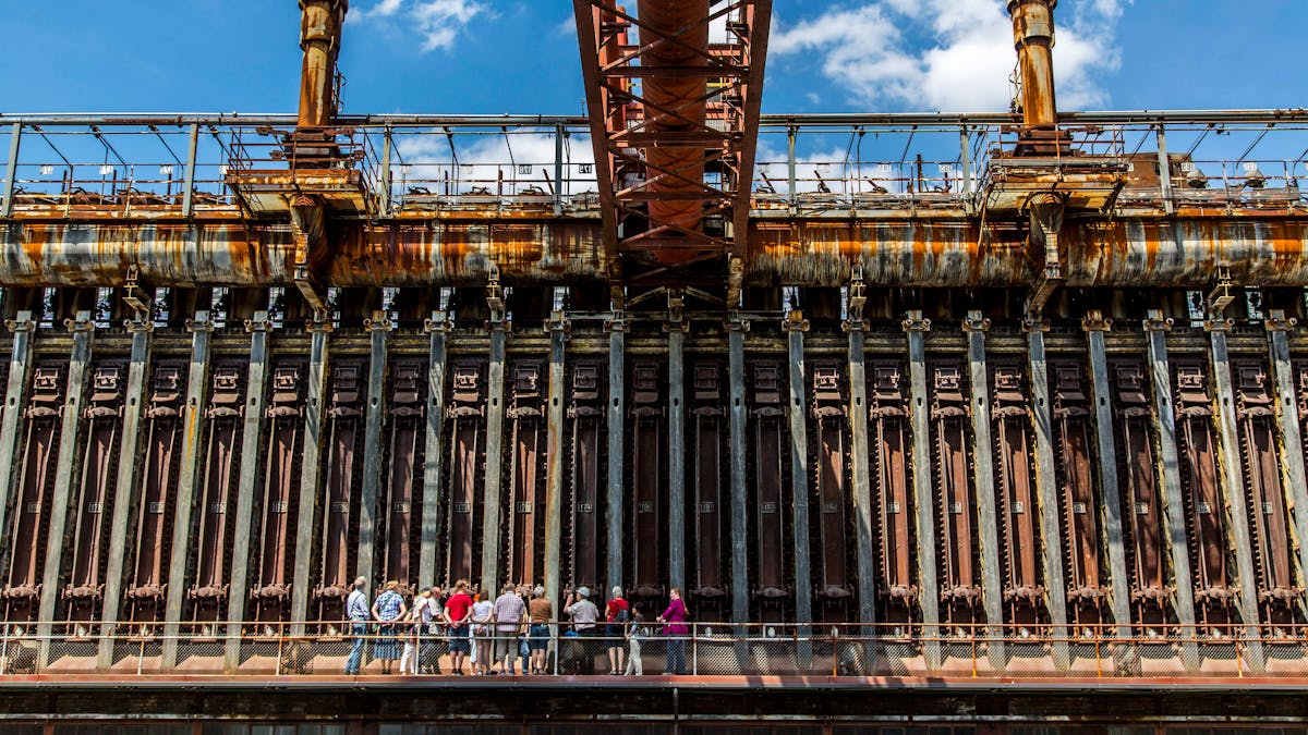 Eine Gruppe von Besuchern steht auf einem schmalen, erhöhten Metallsteg mit Geländer vor einer beeindruckenden, rostigen Industrieanlage. Die massive Wandstruktur besteht aus zahlreichen hohen, dunklen Kammern mit schweren Metalltüren und dicken, vertikalen Verstrebungen. Oberhalb der Anlage verlaufen metallene Rohrleitungen mit sichtbaren Rostspuren, von denen ein großes, rostfarbenes Rohr schräg in die Mitte des Bildes ragt. Darüber befindet sich ein Geländer mit Laufwegen und Plattformen. Die Anlage ist die stillgelegte Kokerei Zollverein, die nun als Industriedenkmal besichtigt werden kann. Die rostige, metallische Oberfläche der Konstruktion kontrastiert mit dem strahlend blauen Himmel mit weißen Wolken im Hintergrund. Die Besucher, einige in farbenfroher Kleidung, betrachten interessiert die monumentale Anlage.