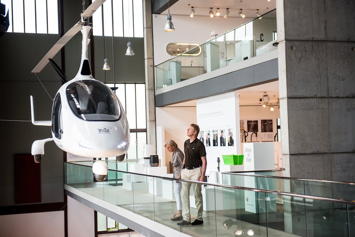 Das Red Dot Design Museum, mit seiner offenen Innenarchitektur und großen Glasflächen, vermittelt einen modernen Eindruck. Im Mittelpunkt des Bildes hängt ein futuristisches, weißes Gyrocopter-Fluggerät mit einer geschlossenen Kabine und einem Rotor oberhalb des Cockpits, scheinbar schwebend an dünnen Seilen befestigt. Zwei Besucher betrachten das Exponat von einem erhöhten Rundgang mit Glasgeländer aus. Im Hintergrund gibt es eine Ausstellung mit verschiedenen modernen Designobjekten, weißen Informationstafeln und beleuchteten Vitrinen. Die Szene vermittelt einen Eindruck von Innovation und technologischem Fortschritt.