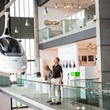 Das Red Dot Design Museum, mit seiner offenen Innenarchitektur und großen Glasflächen, vermittelt einen modernen Eindruck. Im Mittelpunkt des Bildes hängt ein futuristisches, weißes Gyrocopter-Fluggerät mit einer geschlossenen Kabine und einem Rotor oberhalb des Cockpits, scheinbar schwebend an dünnen Seilen befestigt. Zwei Besucher betrachten das Exponat von einem erhöhten Rundgang mit Glasgeländer aus. Im Hintergrund gibt es eine Ausstellung mit verschiedenen modernen Designobjekten, weißen Informationstafeln und beleuchteten Vitrinen. Die Szene vermittelt einen Eindruck von Innovation und technologischem Fortschritt.