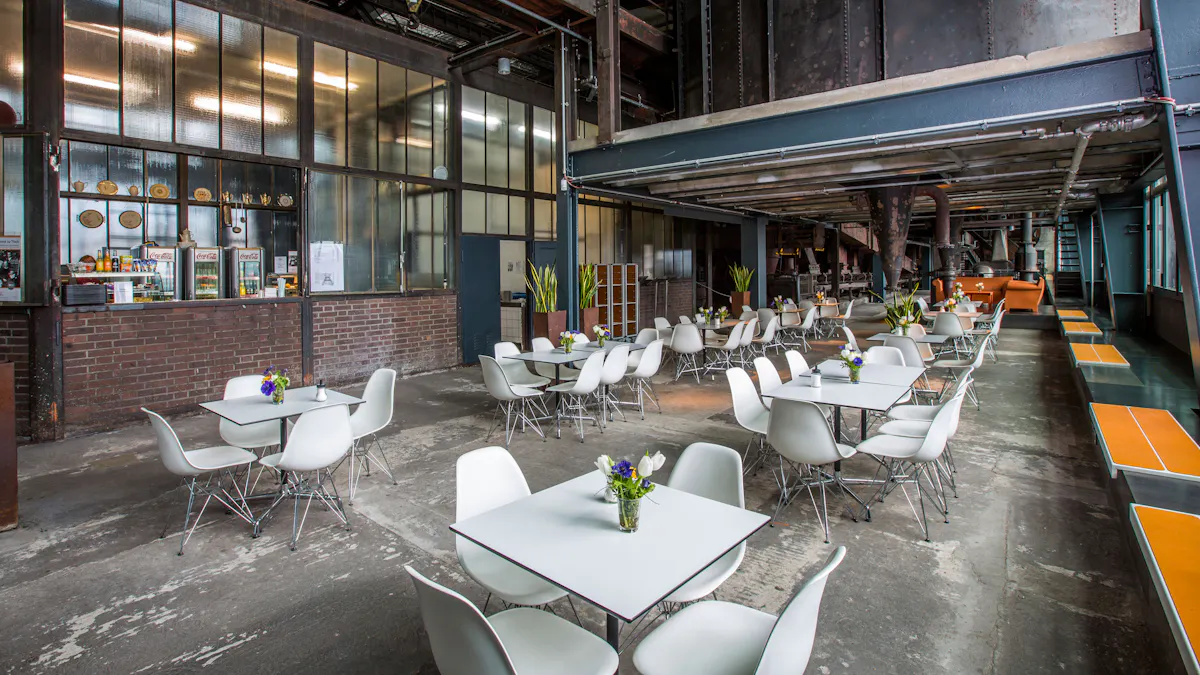 Das Café Kohlenwäsche mit industriellem Flair im Eingangsbereich des Besucherzentrums Ruhr. Die Einrichtung kombiniert weiße, minimalistische Tische und Stühle mit der rauen Ästhetik des alten Industriegebäudes. Die Wände bestehen aus dunklen Backsteinen und großen Fenstern mit Metallrahmen, hinter denen sich ein Selbstbedienungsbereich mit Getränken und Snacks befindet. Der Boden ist aus abgenutztem Beton, was den industriellen Charme unterstreicht. An der rechten Seite gibt es eine lange Sitzbank mit orangefarbenen Flächen, während weiter hinten gemütliche Sitzgruppen mit Pflanzen und Dekorationen für eine einladende Atmosphäre sorgen. Im Hintergrund sind alte Maschinen und Rohrleitungen sichtbar, die die industrielle Vergangenheit des Ortes betonen.