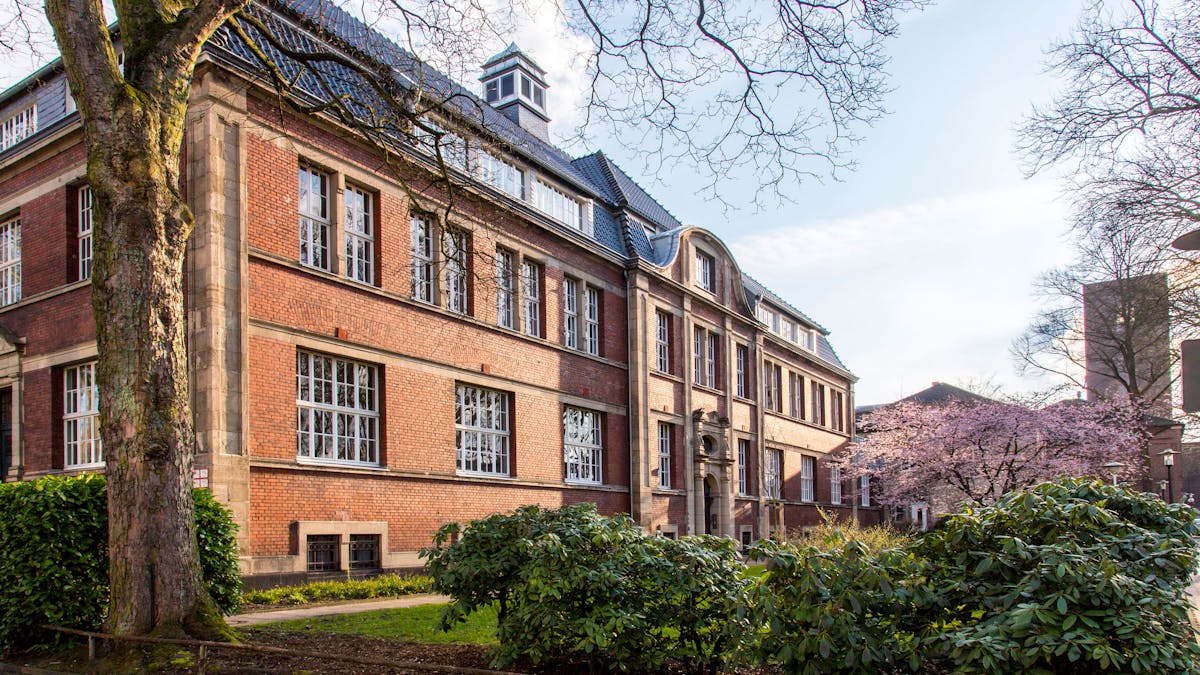 Das Bild zeigt eines der historischen Verwaltungsgebäude der Zeche Zollverein in Essen, ein repräsentativer Bau aus rotem Backstein mit Sandsteinverzierungen. Das Gebäude wurde 1906 errichtet und diente als zentrale Verwaltung der Zeche. Der dreigeschossige Bau besticht durch seine symmetrische Architektur mit großen Sprossenfenstern und einem dunkel gedeckten Walmdach. Über dem Haupteingang ist eine aufwendig gestaltete Sandsteinfassade zu sehen, die den Eingangsbereich betont. An der Dachspitze thront ein kleiner Turm. Im Vordergrund des Bildes sind ein großer Baumstamm sowie grüne Büsche und Beete zu sehen, die Teil der gepflegten Gartenanlage um das Verwaltungsgebäude sind. Rechts im Bild blüht ein Baum mit rosa Blüten. Im Hintergrund sind weitere Gebäude des UNESCO-Welterbes Zollverein zu erkennen, darunter der Förderturm der Schachtanlage 1/2/8.
