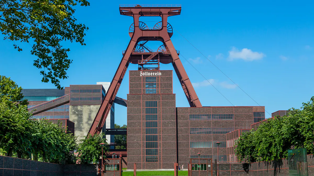 Das Bild zeigt das ikonische Doppelbock-Fördergerüst von Schacht XII auf dem Gelände der Zeche Zollverein in Essen, eingerahmt von Bäumen und blauem Himmel. Das massive Fördergerüst aus Stahl thront über dem Zentralgebäude, das mit seiner dunklen Backsteinfassade und den großen Glasfenstern ein typisches Beispiel für die Neue Sachlichkeit der Industriearchitektur ist. Auf der Fassade des zentralen Gebäudes ist in weißer Schrift das Wort Zollverein zu lesen. Die breiten Förderbänder verbinden die Gebäude miteinander. Die Grünanlagen im Vordergrund bilden einen starken Kontrast zu den rotbraunen, kubischen Gebäuden und verdeutlichen die auf Repräsentation ausgerichtete Architektur dieser einmaligen Industrieanlage.