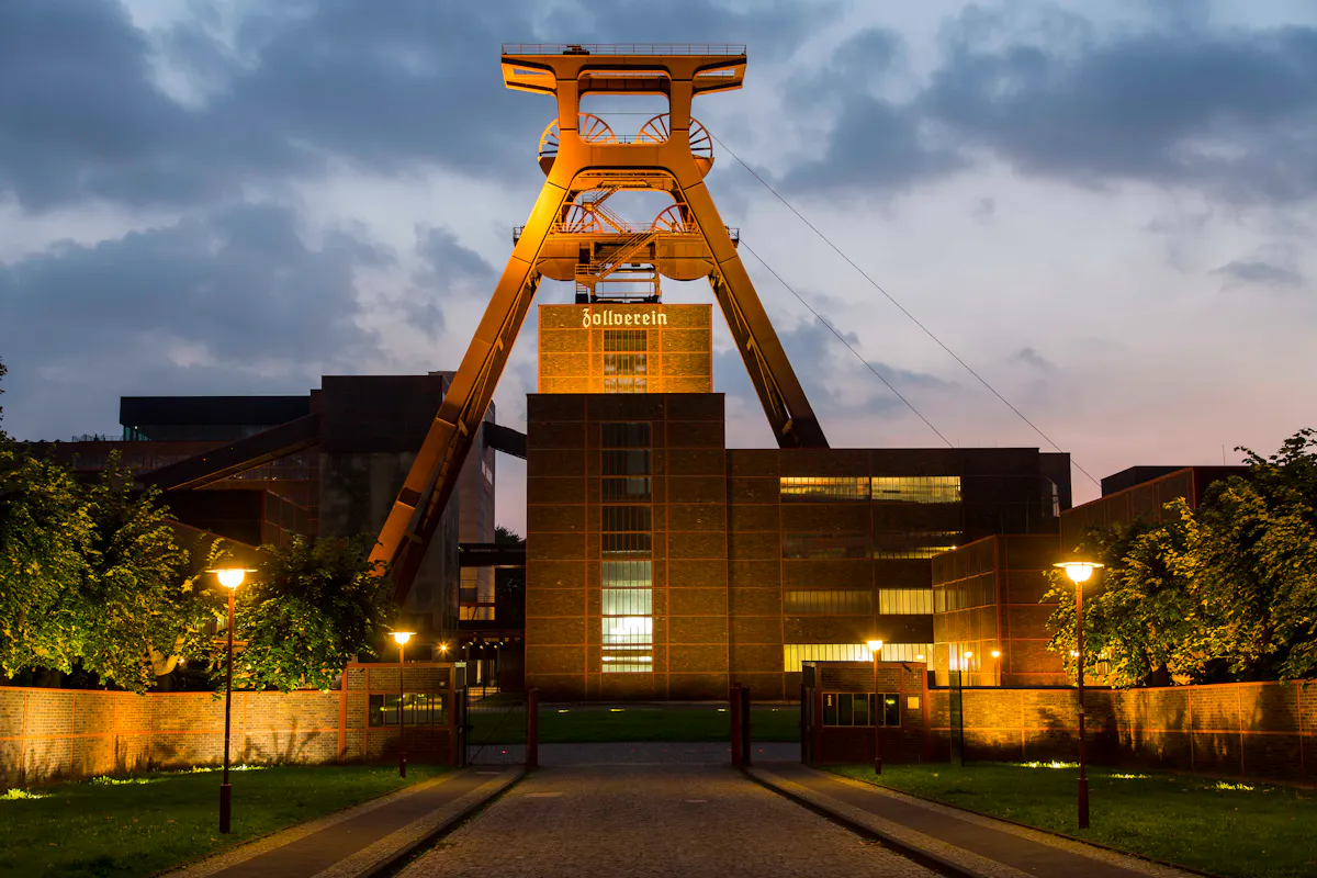 Das beleuchtete Fördergerüst der Zeche Zollverein steht imposant vor einem Abendhimmel mit dunklen Wolken. Die Backsteingebäude des ehemaligen Bergwerks sind ebenfalls von warmem Licht erhellt. Über dem zentralen Gebäude ist der Schriftzug Zollverein zu sehen. Eine gepflasterte Straße führt auf das Gelände zu, flankiert von niedrigen Backsteinmauern, grünen Rasenflächen und mehreren Laternen, die den Weg in sanftem Licht erstrahlen lassen. Die Szenerie verbindet industrielle Architektur mit einer stimmungsvollen Beleuchtung in der Dämmerung.