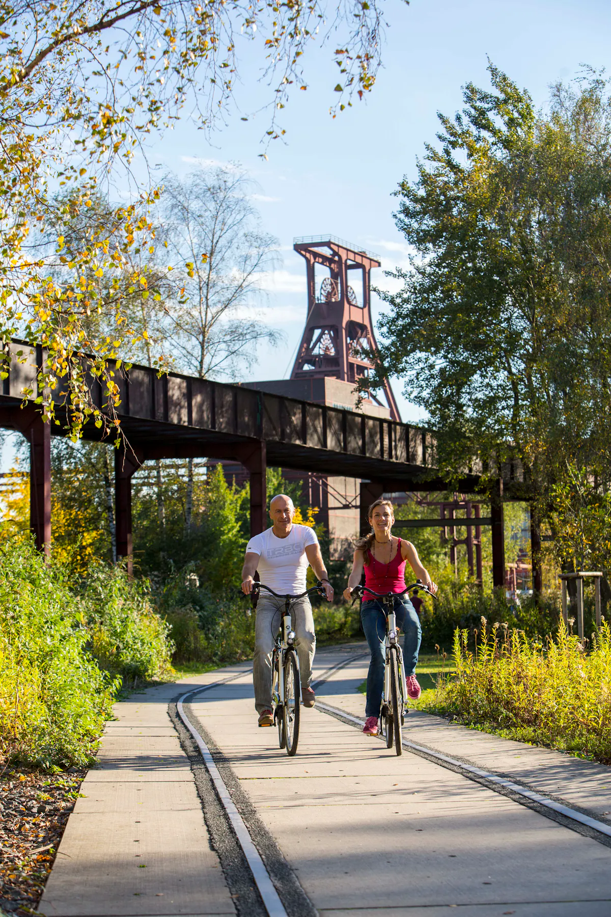 Ein Paar fährt auf Fahrrädern einen asphaltierten Weg entlang, der in eine alte Bahnschiene integriert ist. Der Weg ist von dichtem, grünem Bewuchs und herbstlich gefärbten Bäumen gesäumt und führt durch den Zollverein Park. Im Hintergrund ragt das ikonische rote Fördergerüst der Zeche Zollverein über eine stillgelegte, erhöhte Industriebrücke hinaus. Der Himmel ist klar und blau, und die Sonne wirft lange Schatten, die die warme, herbstliche Atmosphäre verstärken. Die Szene vermittelt einen Eindruck von Freizeit, Natur und Industriekultur in harmonischer Verbindung.