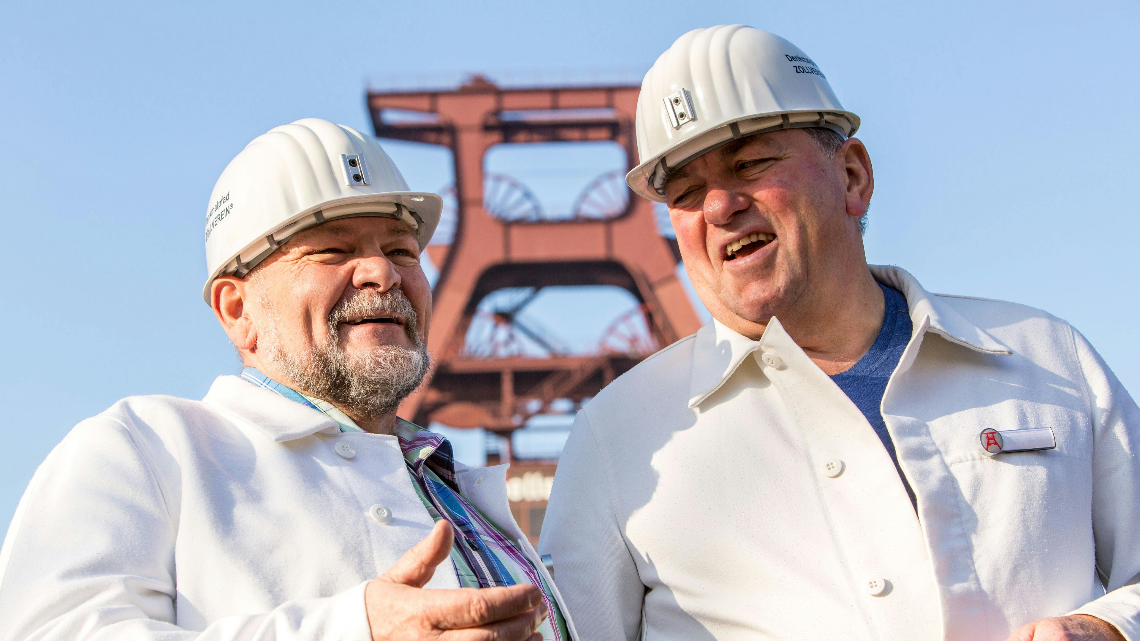 Das Bild zeigt zwei ältere Männer in weißen Bergmannskitteln und Schutzhelmen, die lächelnd vor dem Doppelbock-Fördergerüst der Zeche Zollverein Schacht XII stehen und sich angeregt unterhalten. Die Schutzhelme tragen das Logo von Zollverein, und einer der Männer hat ein Namensschild mit dem stilisierten Fördergerüst an seiner Jacke. Dies zeigt, dass sie mit der Geschichte der Zeche eng verbunden sind und sie heutzutage Führungen anbieten. Im Hintergrund ragt das rote Fördergerüst von Schacht XII in den blauen Himmel – das Wahrzeichen des Welterbes.
