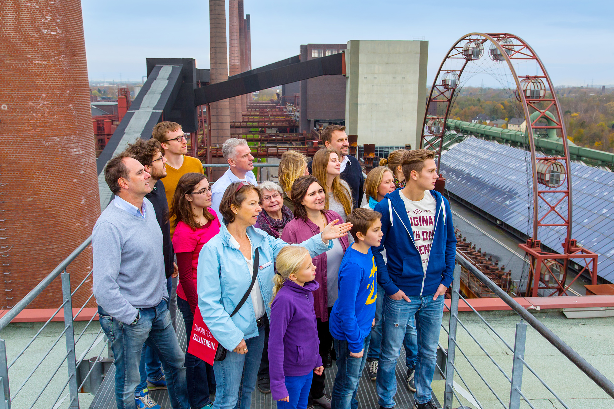 Eine Gruppe von Menschen, darunter Erwachsene und Kinder, steht auf einer Aussichtsplattform auf der Mischanlage der Kokerei Zollverein. Die Personen schauen interessiert in eine Richtung, während eine Frau in einer hellblauen Jacke mit einer roten Denkmalpfad Zollverein-Tasche etwas erklärt. Im Hintergrund sind große Backsteinschornsteine, metallene Industriebauten und ein auffälliges, rostiges Riesenrad mit geschlossenen Gondeln zu sehen. Eine lange Reihe von Solarpanelen erstreckt sich auf einem Dach, was auf die nachhaltige Nutzung des Areals hinweist.