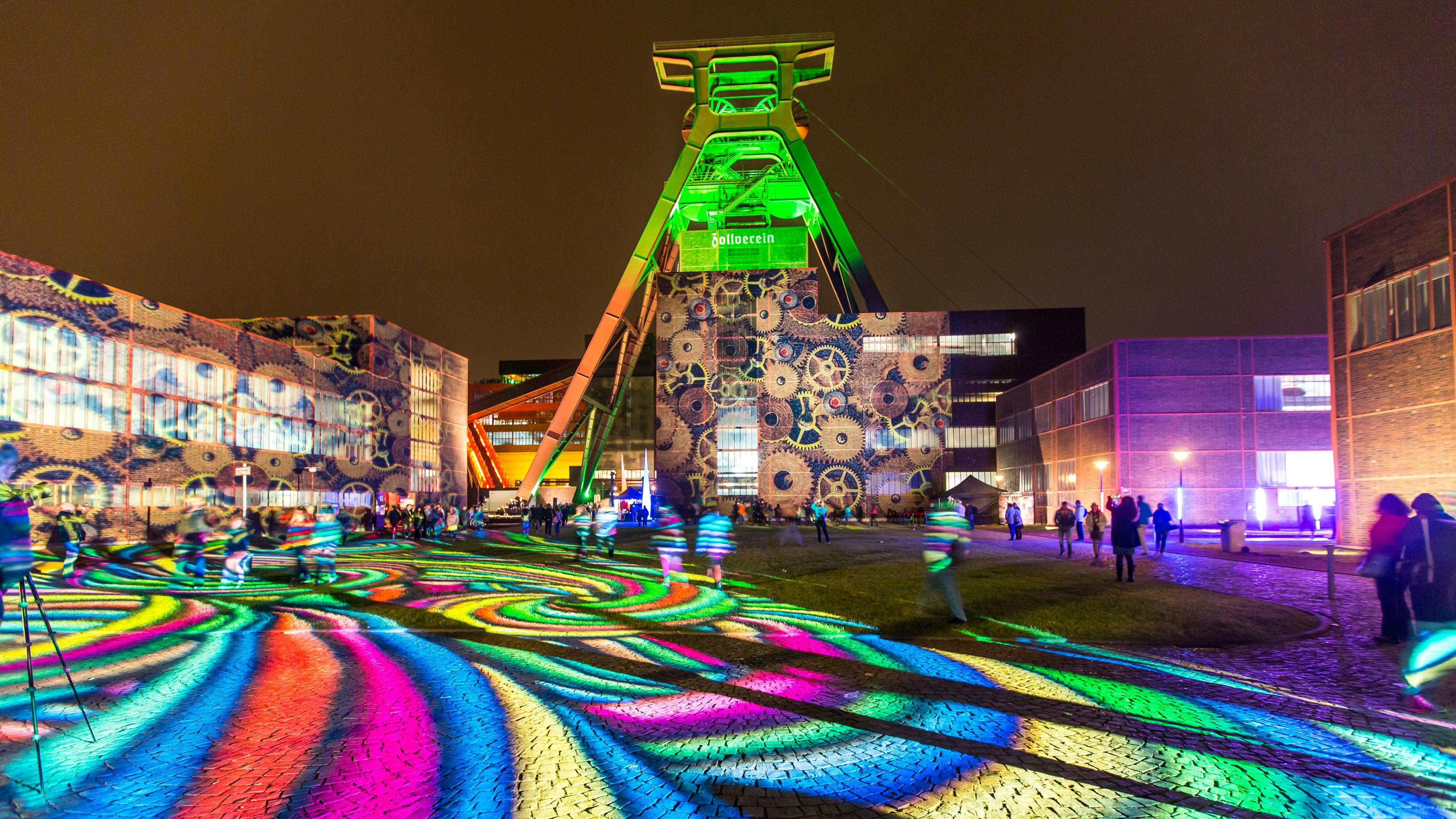 Eine beeindruckende Lichtinstallation, anlässlich der jährlich stattfindenden Extraschicht, verwandelt das Gelände der Zeche Zollverein in ein farbenfrohes Kunstwerk bei Nacht. Das große Fördergerüst ist in leuchtendem Grün angestrahlt, während auf die Backsteinfassade darunter ein Muster aus ineinandergreifenden Zahnrädern projiziert wird. Der gepflasterte Boden ist mit bunten, wirbelnden Lichtmustern in Rot, Blau, Gelb, Grün und Lila überzogen, die sich in die Umgebung ausbreiten. Mehrere Personen laufen oder stehen auf dem Platz, einige sind mit Kameras oder Stativen unterwegs.