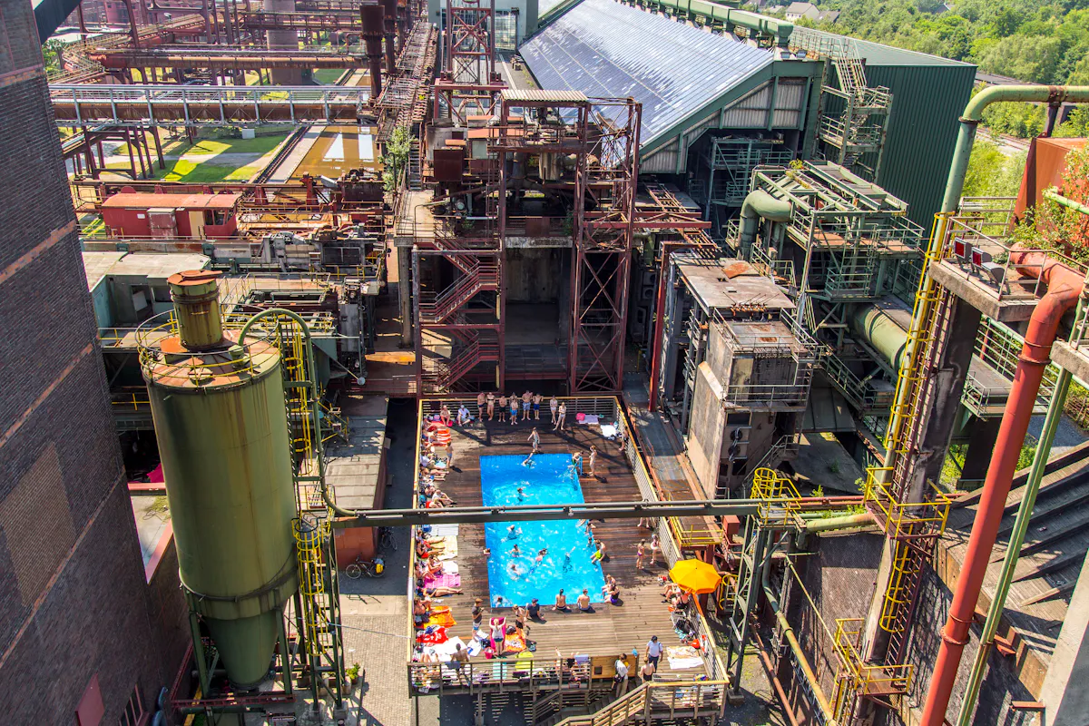 Das Kunstprojekt Werksschwimmbad auf der Kokerei Zollverein ist während der Sommerferien ein ungewöhnliches Freibad inmitten einer stillgelegten Industrieanlage. In der Mitte des Bildes befindet sich ein rechteckiger, leuchtend blauer Pool, umgeben von einem hölzernen Sonnendeck mit Liegestühlen, Handtüchern und einem orangefarbenen Sonnenschirm. Mehrere Menschen baden oder entspannen sich am Beckenrand. Das gesamte Areal ist umgeben von alten, rostigen Metallstrukturen, großen Rohren, Treppen und Industriebauten mit bunten Stahlgerüsten. Im Hintergrund sind weitere Rohrleitungen, Maschinenanlagen und ein Gebäude mit einem großen Dach voller Solarpaneele zu sehen. Die Szene zeigt die kreative Umnutzung eines ehemaligen Industriekomplexes in einen Freizeitort mit besonderem Charme.