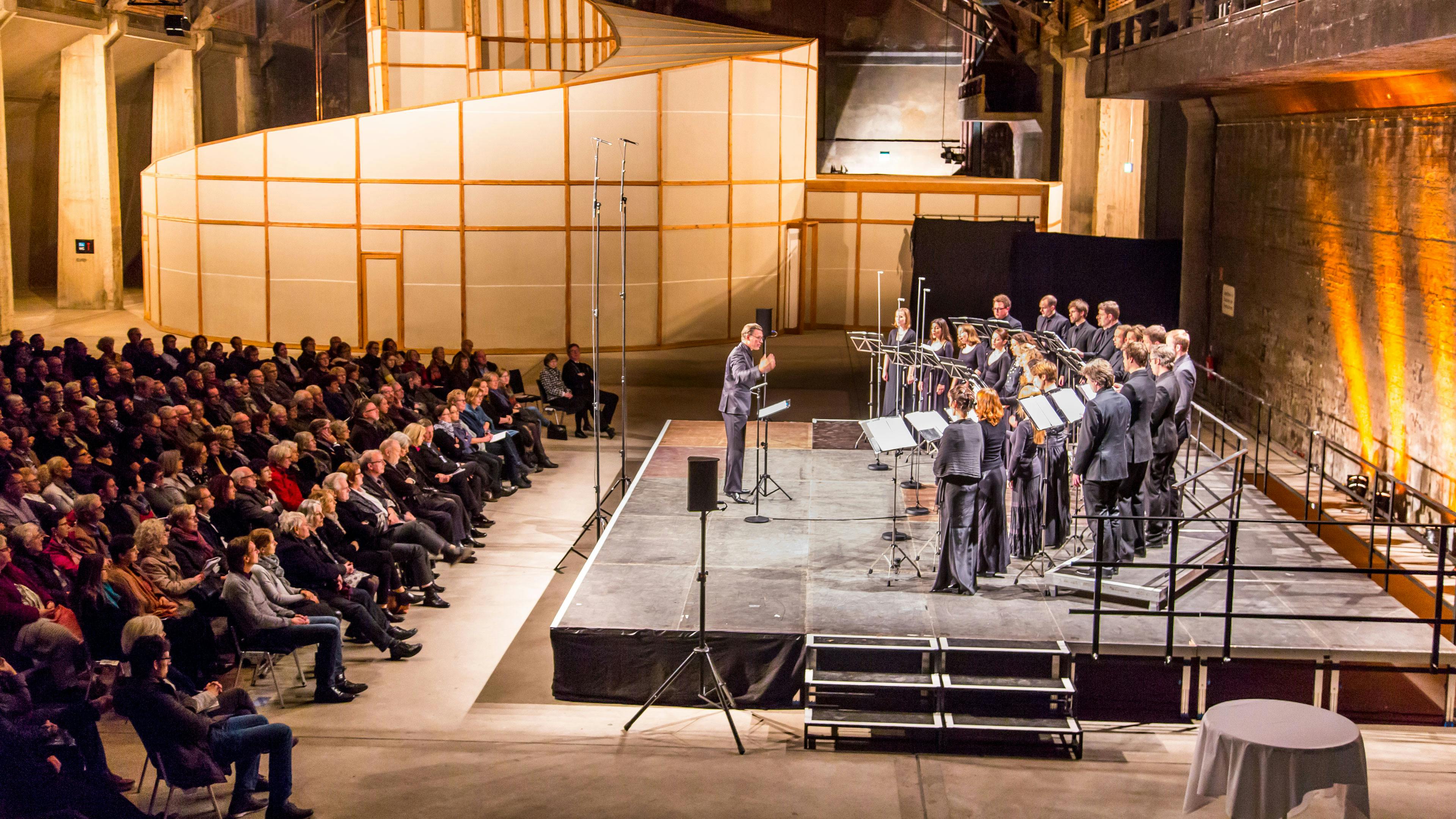 Das Salzlager auf der Kokerei Zollverein als stilvoll beleuchteter Konzertsaal mit industriellem Flair, in dem ein Chor auf einer erhöhten Bühne auftritt. Die Sängerinnen und Sänger, alle in eleganter schwarzer Kleidung, stehen in Reihen hinter Notenständern, während ein Dirigent vor ihnen steht und den Takt angibt. Mikrofonständer sind auf der Bühne verteilt, und Lautsprecher stehen am Rand. Das Publikum sitzt aufmerksam in mehreren Reihen von Stühlen, in dunkler Kleidung, mit teils verschränkten Armen oder Händen im Schoß. Der Raum hat eine rohe, industrielle Ästhetik mit Betonwänden, die in warmem, orangefarbenem Licht angestrahlt werden. Im Hintergrund befindet sich eine große, schneckenhausförmige Struktur aus Holzrahmen und hellen Stoffpaneelen, die hier als dekoratives Element dient. Letzteres ist eigentlich das begehbare Kunstwerk Palace of Projects, welches an Wochenenden besichtigt werden kann.