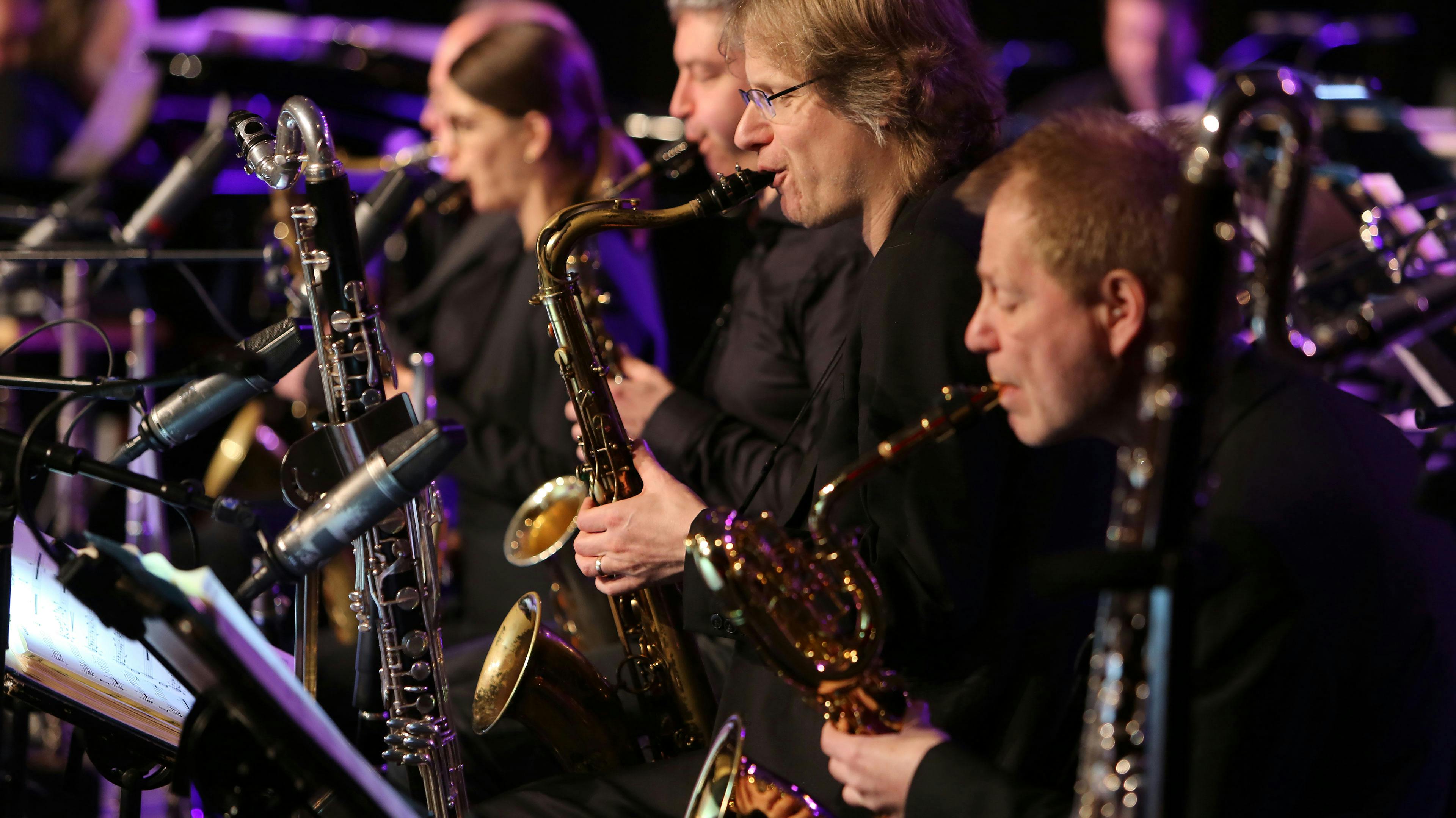 Eine Bläsersektion spielt konzentriert während eines Konzerts auf dem UNESCO-Welterbe Zollverein. Mehrere Musiker in schwarzer Kleidung sitzen in einer Reihe und spielen Saxophone und Klarinetten. Ihre Instrumente glänzen im warmen Bühnenlicht, während Mikrofone nah an den Schalltrichtern positioniert sind, um den Klang einzufangen. Notenblätter auf Pulten vor den Musikern sind sichtbar, deuten aber nur schemenhaft ihre Inhalte an. Die Bühne ist in stimmungsvolles, violett-blaues Licht getaucht, was eine atmosphärische, fast jazzige Konzertstimmung vermittelt. Das Bild zeigt die hohe musikalische Qualität und die besondere Atmosphäre der Zollverein Konzerte, die regelmäßig auf dem ehemaligen Industriegelände stattfinden und das denkmalgeschützte Ambiente der Zeche mit erstklassiger Livemusik verbinden.