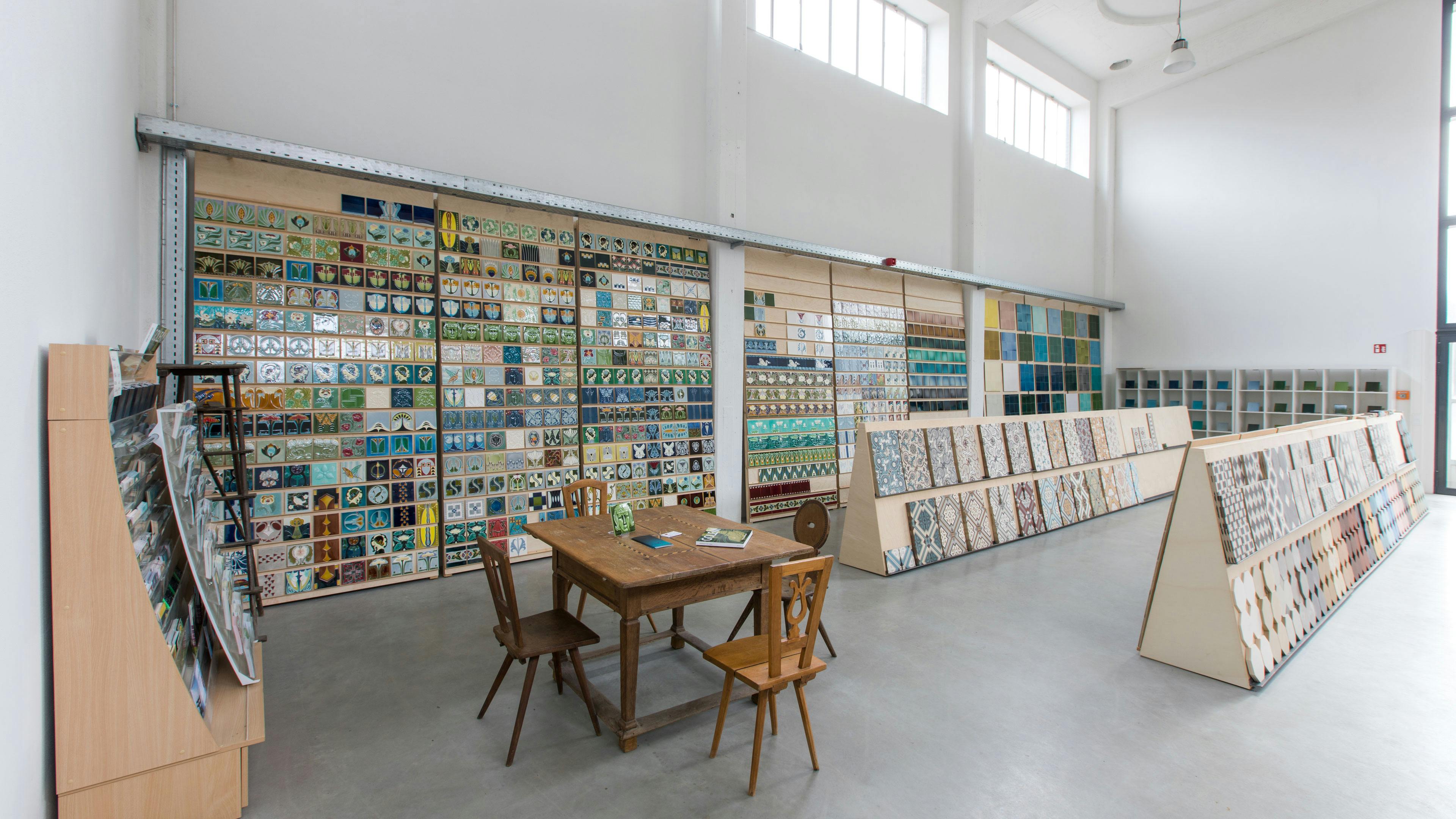 Ein heller, geräumiger Ausstellungsraum mit hohen Decken und großen Fenstern, der eine breite Auswahl an dekorativen Fliesen präsentiert. An der linken Wand sind zahlreiche kunstvoll gestaltete Fliesen in verschiedenen Farben, Mustern und Motiven angebracht, vor allem in Blau- und Grüntönen mit floralen und geometrischen Designs. In der Mitte des Raumes stehen mehrere schräg aufgestellte Holztafeln mit einer Vielzahl von Fliesenmustern, die von klassischen bis zu modernen Designs reichen. Im Vordergrund befindet sich ein rustikaler Holztisch mit drei unterschiedlich gestalteten Holzstühlen, auf dem einige Musterbücher oder Kataloge liegen. Die Atmosphäre ist minimalistisch und ordentlich, mit einem Betonboden und weißen Wänden, die den Fokus auf die Fliesenausstellung lenken.