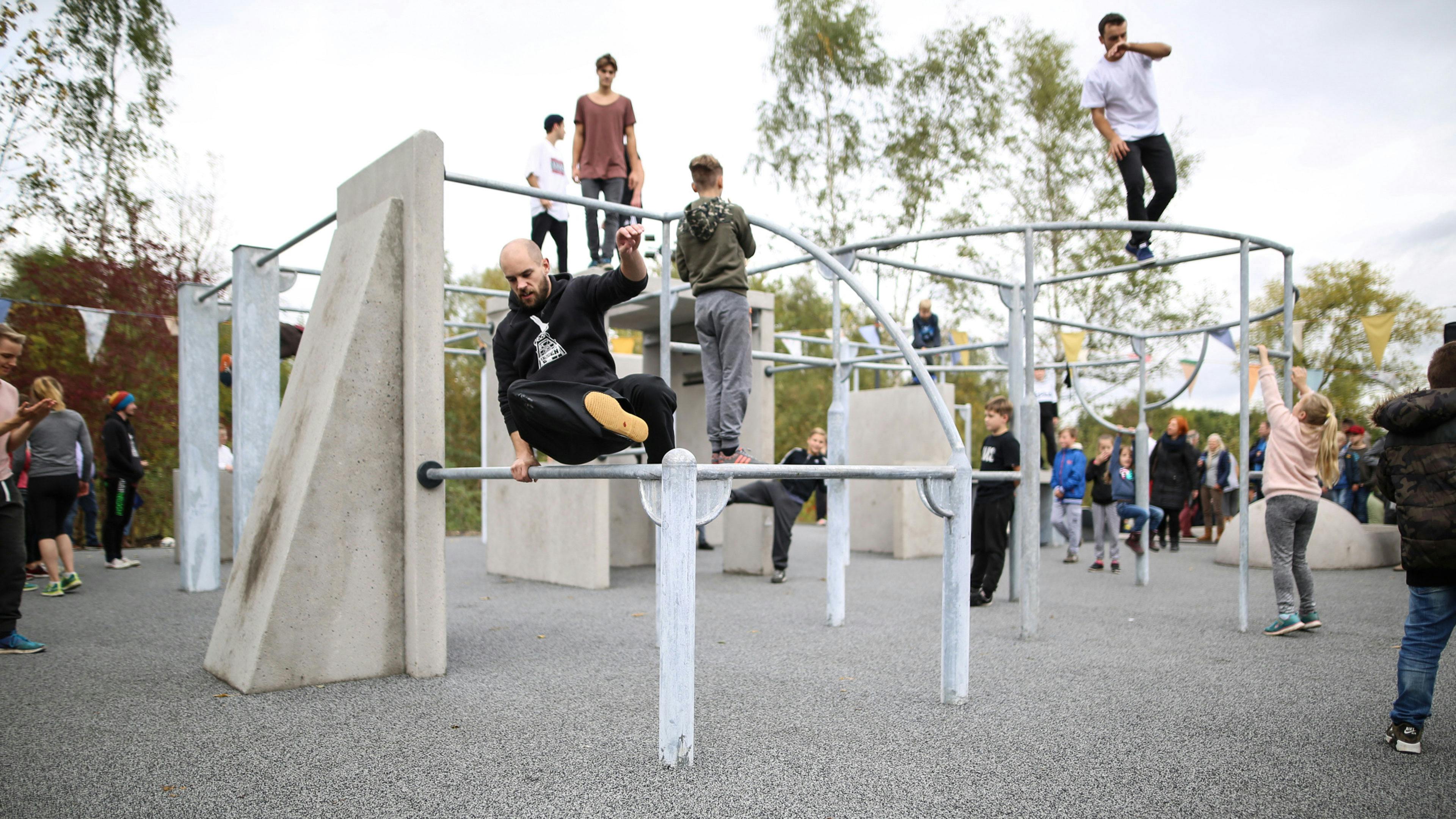 Das Bild zeigt eine Parkour-Anlage auf dem Gelände des UNESCO-Welterbes Zollverein in Essen, die von Menschen unterschiedlichen Alters genutzt wird. Im Vordergrund springt ein Mann mit einem schwarzen Hoodie über eine Metallstange, während er sich mit einer Hand festhält. Rechts daneben klettert ein Mädchen in einer rosa Jacke an einer Metallkonstruktion hoch. Die Anlage besteht aus verschiedenen Beton- und Metallstrukturen, darunter schräge Wände, Stangen, Klettergerüste und Plattformen. Auf und zwischen den Hindernissen bewegen sich Kinder, Jugendliche und Erwachsene, die Parkour- oder Kletterübungen ausführen. Im Hintergrund beobachten Zuschauer das Geschehen, einige tragen Sportkleidung, andere sind als Besucher vor Ort. Der Bereich ist von Bäumen und Sträuchern umgeben und bunte Wimpel hängen in der Luft. Die moderne Parkour-Anlage ist Teil der Freizeit- und Sportangebote auf Zollverein, das sich als Kultur- und Erlebnisstandort weiterentwickelt hat.