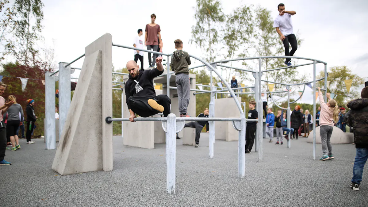 Das Bild zeigt eine Parkour-Anlage auf dem Gelände des UNESCO-Welterbes Zollverein in Essen, die von Menschen unterschiedlichen Alters genutzt wird. Im Vordergrund springt ein Mann mit einem schwarzen Hoodie über eine Metallstange, während er sich mit einer Hand festhält. Rechts daneben klettert ein Mädchen in einer rosa Jacke an einer Metallkonstruktion hoch. Die Anlage besteht aus verschiedenen Beton- und Metallstrukturen, darunter schräge Wände, Stangen, Klettergerüste und Plattformen. Auf und zwischen den Hindernissen bewegen sich Kinder, Jugendliche und Erwachsene, die Parkour- oder Kletterübungen ausführen. Im Hintergrund beobachten Zuschauer das Geschehen, einige tragen Sportkleidung, andere sind als Besucher vor Ort. Der Bereich ist von Bäumen und Sträuchern umgeben und bunte Wimpel hängen in der Luft. Die moderne Parkour-Anlage ist Teil der Freizeit- und Sportangebote auf Zollverein, das sich als Kultur- und Erlebnisstandort weiterentwickelt hat.