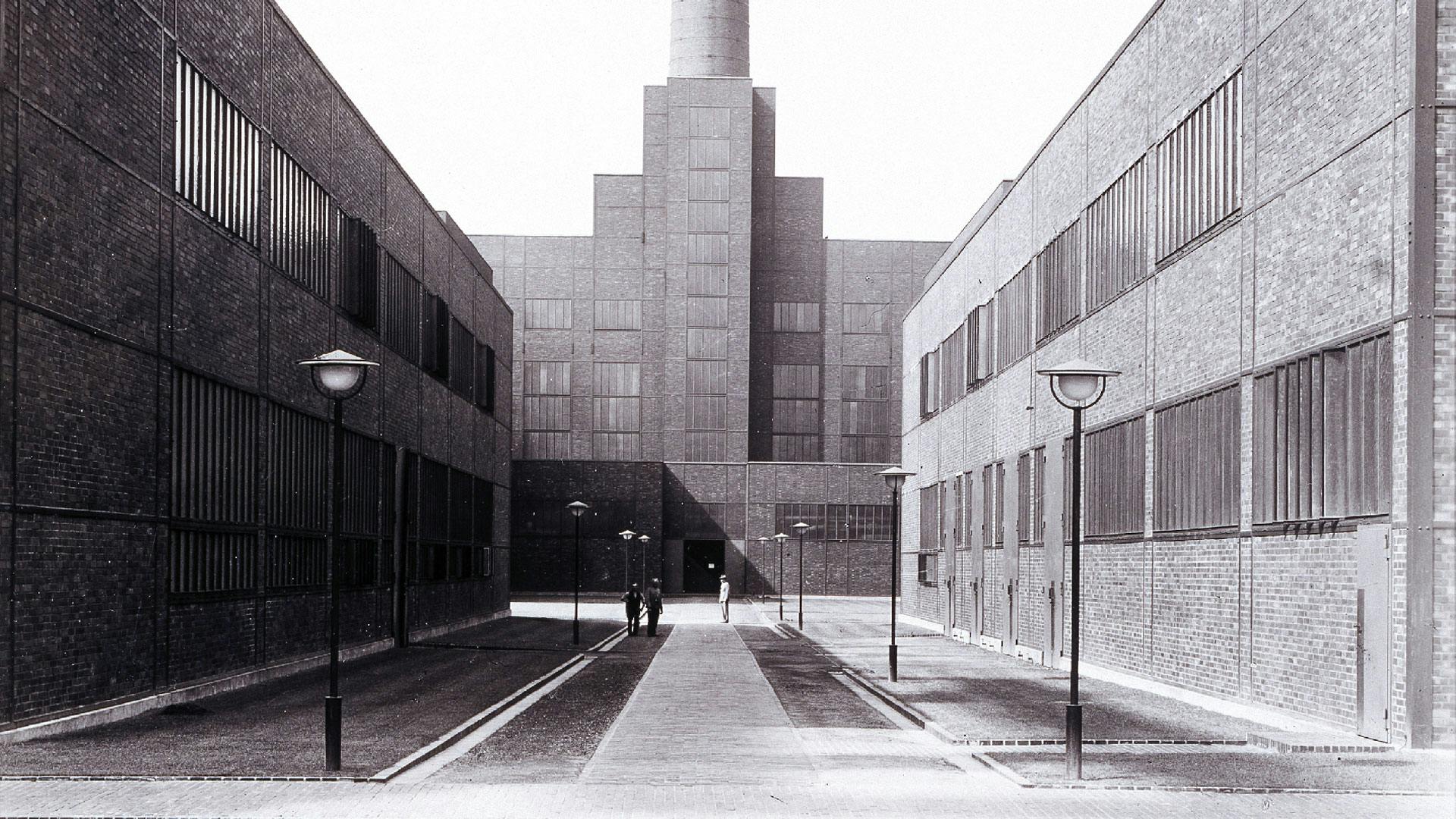 Das Bild zeigt eine historische Schwarz-Weiß-Aufnahme des Kesselhauses der Zeche Zollverein Schacht XII in Essen. Das Kesselhaus ist das zentrale Gebäude im Hintergrund der Aufnahme und zeichnet sich durch seine monumentale Backsteinfassade mit einer gestuften Architektur aus. Ein hoher Schornstein ragt aus dem Bau empor, was auf seine Funktion als Energiezentrale der Zeche hinweist. Der Zugang zum Kesselhaus führt durch einen symmetrisch gestalteten Innenhof, der von zwei langgezogenen Backsteingebäuden mit schmalen, vertikal ausgerichteten Fensterbändern gesäumt wird. Diese flankierenden Gebäude beherbergten einst Werkstätten der Zeche. Ein gepflasterter Weg mit beidseitigen Straßenlaternen führt direkt auf das Kesselhaus zu. Drei Personen sind im Innenhof unterwegs: Zwei gehen gemeinsam, während eine einzelne Person sich von der Tür des Kesselhauses entfernt. Die Architektur spiegelt den Stil der Neuen Sachlichkeit wider, den die Architekten Fritz Schupp und Martin Kremmer bei der Gestaltung von Zollverein XII konsequent verfolgten. Die klaren Linien, reduzierten Verzierungen und die funktionale Anordnung der Gebäude machten die Anlage zu einer der modernsten Zechen ihrer Zeit.