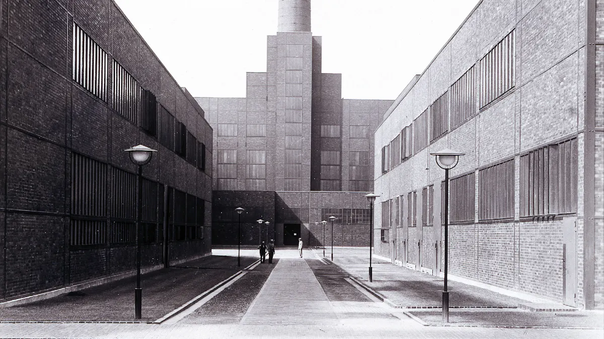 Das Bild zeigt eine historische Schwarz-Weiß-Aufnahme des Kesselhauses der Zeche Zollverein Schacht XII in Essen. Das Kesselhaus ist das zentrale Gebäude im Hintergrund der Aufnahme und zeichnet sich durch seine monumentale Backsteinfassade mit einer gestuften Architektur aus. Ein hoher Schornstein ragt aus dem Bau empor, was auf seine Funktion als Energiezentrale der Zeche hinweist. Der Zugang zum Kesselhaus führt durch einen symmetrisch gestalteten Innenhof, der von zwei langgezogenen Backsteingebäuden mit schmalen, vertikal ausgerichteten Fensterbändern gesäumt wird. Diese flankierenden Gebäude beherbergten einst Werkstätten der Zeche. Ein gepflasterter Weg mit beidseitigen Straßenlaternen führt direkt auf das Kesselhaus zu. Drei Personen sind im Innenhof unterwegs: Zwei gehen gemeinsam, während eine einzelne Person sich von der Tür des Kesselhauses entfernt. Die Architektur spiegelt den Stil der Neuen Sachlichkeit wider, den die Architekten Fritz Schupp und Martin Kremmer bei der Gestaltung von Zollverein XII konsequent verfolgten. Die klaren Linien, reduzierten Verzierungen und die funktionale Anordnung der Gebäude machten die Anlage zu einer der modernsten Zechen ihrer Zeit.