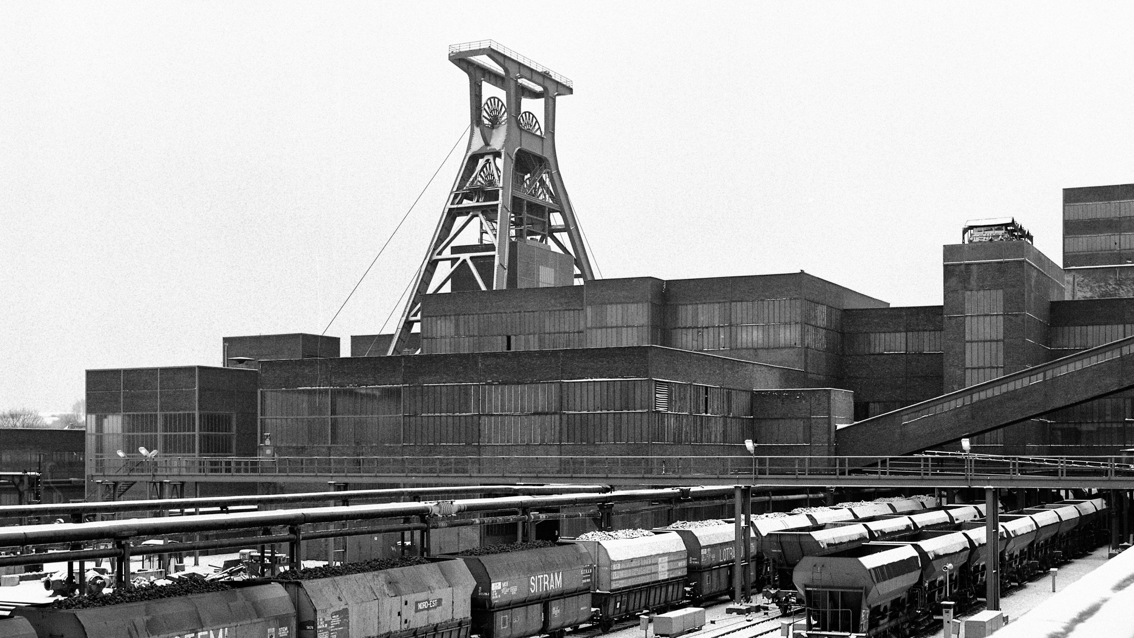 Das Bild zeigt eine historische Schwarz-Weiß-Aufnahme der Zeche Zollverein Schacht XII in Essen. Im Zentrum steht das ikonische Doppelbock-Fördergerüst, das mit seinen großen Seilscheiben und der klaren Stahlkonstruktion über den umliegenden Gebäuden thront. Die Architektur der Zeche ist geprägt von funktionaler, geometrischer Formensprache mit klaren Linien. Die langgezogenen Backsteinfassaden der Kohlenwäsche, der Aufbereitung und der Maschinenhäuser sind mit großen Fensterflächen durchzogen. Im Vordergrund sind mehrere mit Kohle beladene Güterwaggons zu sehen, die auf Schienen an der Verladeanlage stehen. Große Rohre verlaufen horizontal über der Bahnstrecke und verbinden verschiedene Anlagenteile miteinander. Die winterliche Szenerie mit Schnee auf den Waggons und Gebäuden unterstreicht die Atmosphäre des industriellen Ruhrgebiets zur Betriebszeit der Zeche.