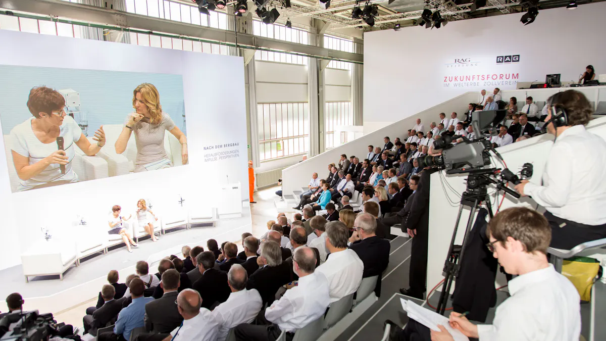 Das Bild zeigt eine Veranstaltung des Zukunftsforums RAG im Welterbe Zollverein. In einem modernen, hellen Konferenzraum sitzen zahlreiche Teilnehmende auf einer ansteigenden Tribüne. Viele von ihnen tragen weiße Hemden und dunkle Anzüge. Im Vordergrund filmt ein Kameramann mit professionellem Equipment, während eine weitere Person Notizen macht. Auf der Bühne führen zwei Frauen ein Gespräch, das live auf eine riesige Leinwand projiziert wird. Die Leinwand zeigt außerdem die Aufschrift: Nach dem Bergbau – Herausforderungen, Impulse, Perspektiven. Im Hintergrund steht eine Person in einem auffälligen orangefarbenen Bergbau-Schutzanzug. Das Zukunftsforum wird von der RAG-Stiftung organisiert und diskutiert Perspektiven für die Zeit nach dem Steinkohlenbergbau.