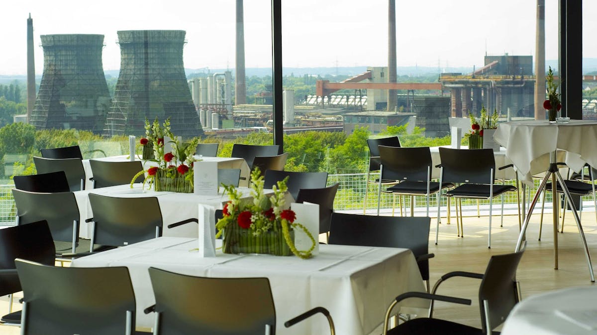 Ein stilvoll eingerichteter Veranstaltungsraum mit großen Glasfenstern, die einen Panoramablick auf die ehemalige Kokerei Zollverein mit zwei markanten Kühltürmen und mehreren Schornsteinen ermöglichen. Die Tische sind mit weißen Tischdecken, schwarzen Stühlen und eleganten Blumenarrangements in Grün, Weiß und Rot dekoriert. Zusätzlich stehen einige hohe Stehtische mit weißen Hussen und schlichten Blumengestecken bereit. Die Szenerie, im Erich-Brost-Pavillon auf dem Dach der Kohlenwäsche aufgenommen, verbindet modernes Design mit industriellem Charme und einem Blick ins Grüne.