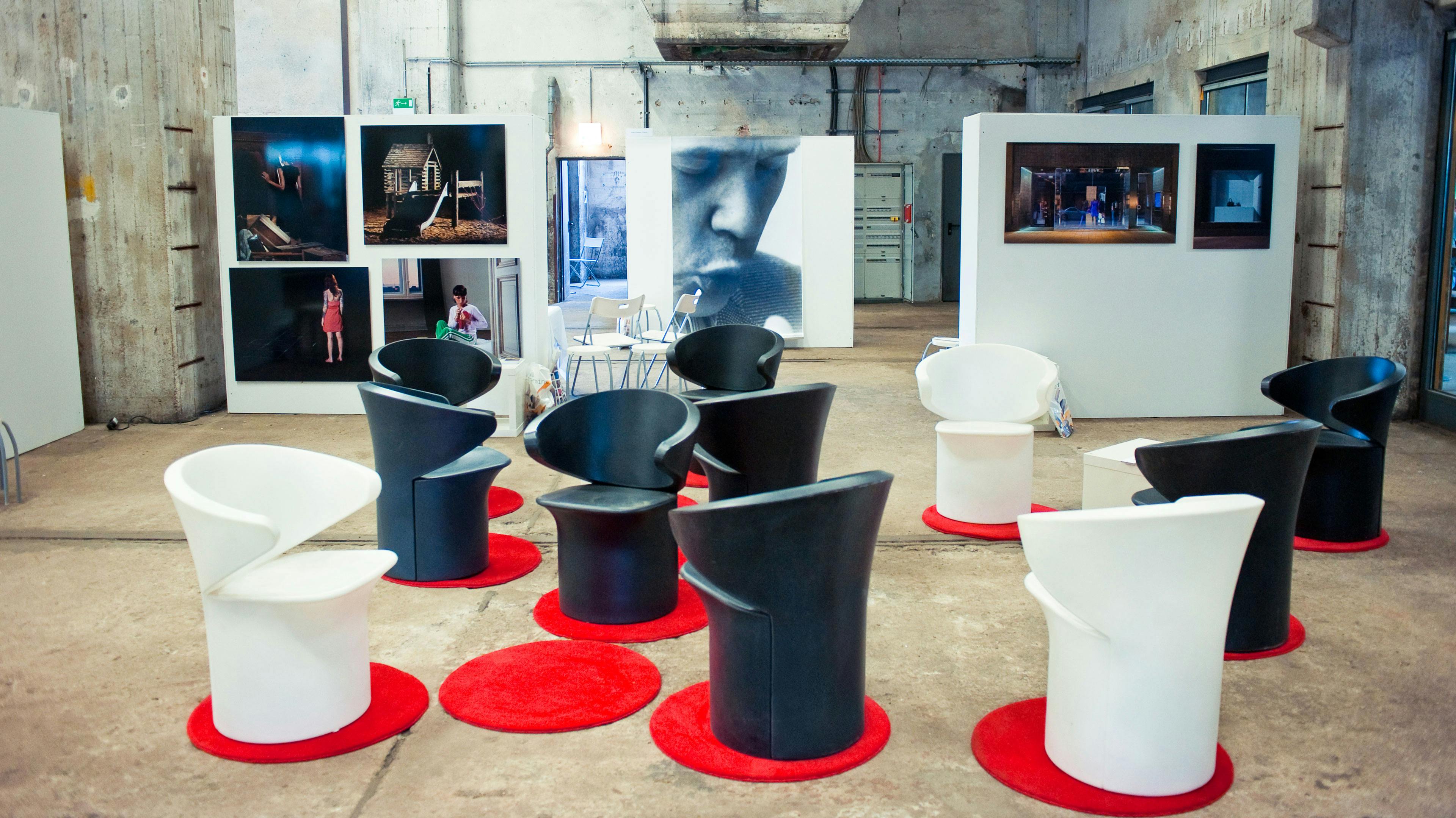 Ein künstlerisch gestalteter Veranstaltungsraum mit einer Sitzgruppe aus schwarzen und weißen Designstühlen, die auf kleinen, runden, roten Teppichen angeordnet sind. Die Stühle haben eine moderne, geschwungene Form. Der Boden besteht aus rohem Beton, und die Wände haben eine industrielle, unverputzte Optik mit sichtbaren Betonstrukturen und Metallrohren. Im Hintergrund sind große weiße Stellwände mit verschiedenen Fotografien zu sehen, darunter Porträtaufnahmen und künstlerische Szenen. Eine große Schwarz-Weiß-Fotografie eines nachdenklich blickenden Mannes dominiert die Mitte des Hintergrunds. Auf der linken Seite sind einige Klappstühle und eine offene Tür zu erkennen, die in einen weiteren Bereich führt.