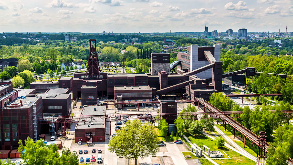 Das Bild zeigt eine Luftaufnahme der Zeche Zollverein in Essen, eine der bekanntesten Industriedenkmäler Europas. Im Zentrum des Bildes ragt das markante Doppelbock-Fördergerüst von Schacht 12 auf, das als Wahrzeichen des Ruhrgebiets gilt. Die umliegenden Backsteinbauten mit ihren typischen Fensterbändern gehören zu den historischen Förderanlagen, Maschinenhallen und Werkstätten der ehemaligen Zeche Zollverein. Lange, schräge Förderbandbrücken verbinden die einzelnen Gebäudekomplexe und führen auch zur Kohlenwäsche, dem heutigen Standort des Ruhr Museums. Im Vordergrund sind rote Stahlrohrleitungen und Gehwege des renaturierten Zechengeländes zu sehen, das heute als Kultur- und Freizeitort dient. Auf der rechten Bildseite erstreckt sich eine begrünte Landschaft. Im Hintergrund ist die Skyline der Stadt Essen mit Hochhäusern, Kirchtürmen und dem markanten Fernsehturm sichtbar.