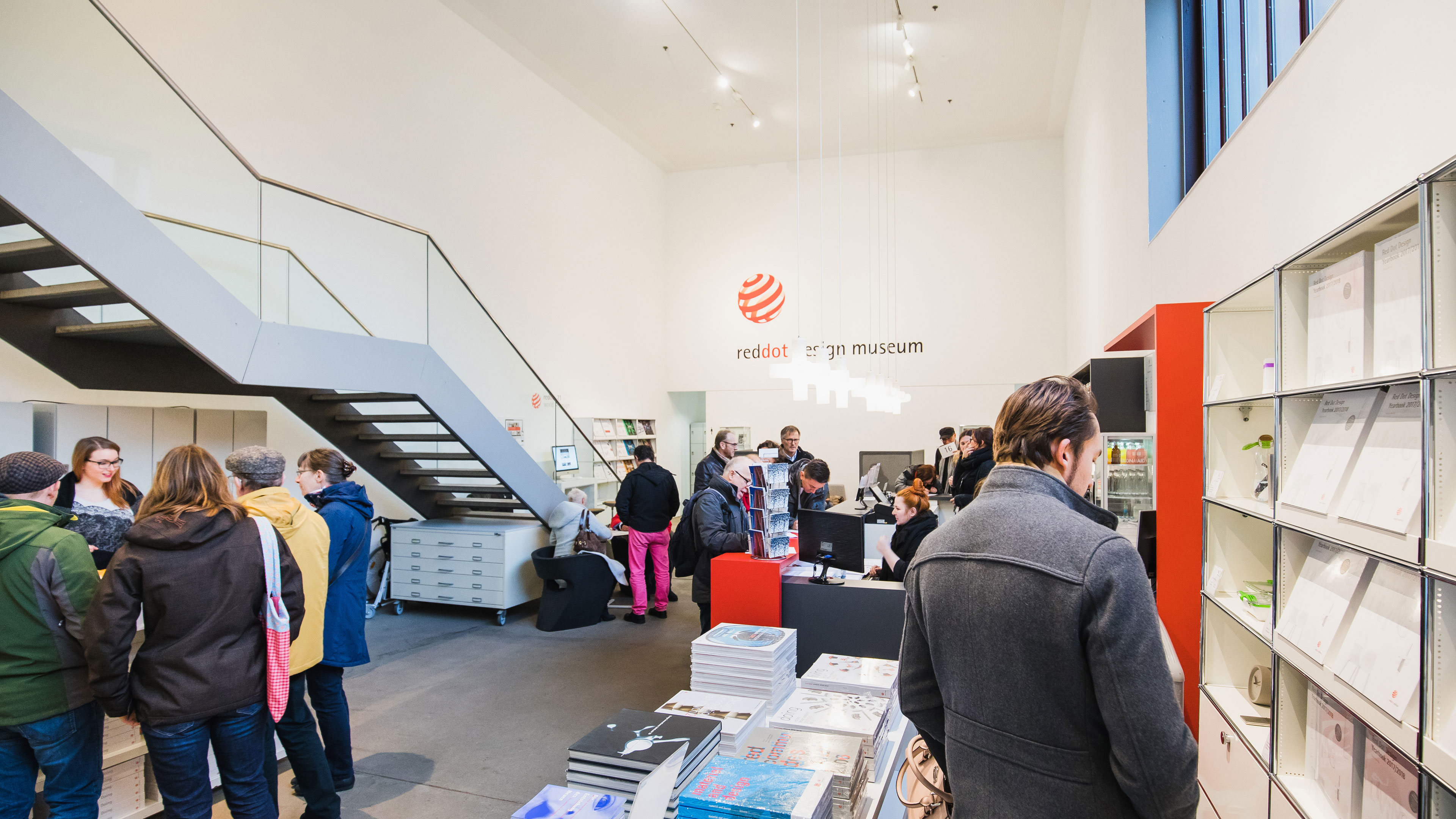 Ein belebter Eingangsbereich des Red Dot Design Museums mit modernen, weißen Wänden und einer offenen Treppe mit Glasgeländer auf der linken Seite. Über dem Empfangsbereich hängt das rote Logo des Museums mit der Aufschrift Red Dot Design Museum. Mehrere Besucher stehen an der Rezeption an, wo eine Mitarbeiterin hinter einem roten Tresen Auskünfte gibt. Andere Gäste stöbern in Designbüchern und Magazinen, die in einem Regal auf der rechten Seite und auf einem Tisch in der Mitte des Raums ausliegen. Im Hintergrund unterhalten sich weitere Besucher oder erkunden den Shopping-Bereich. Die Atmosphäre ist modern und einladend, mit hellen Lichtinstallationen, minimalistischer Möblierung und Designobjekten in den Regalen.