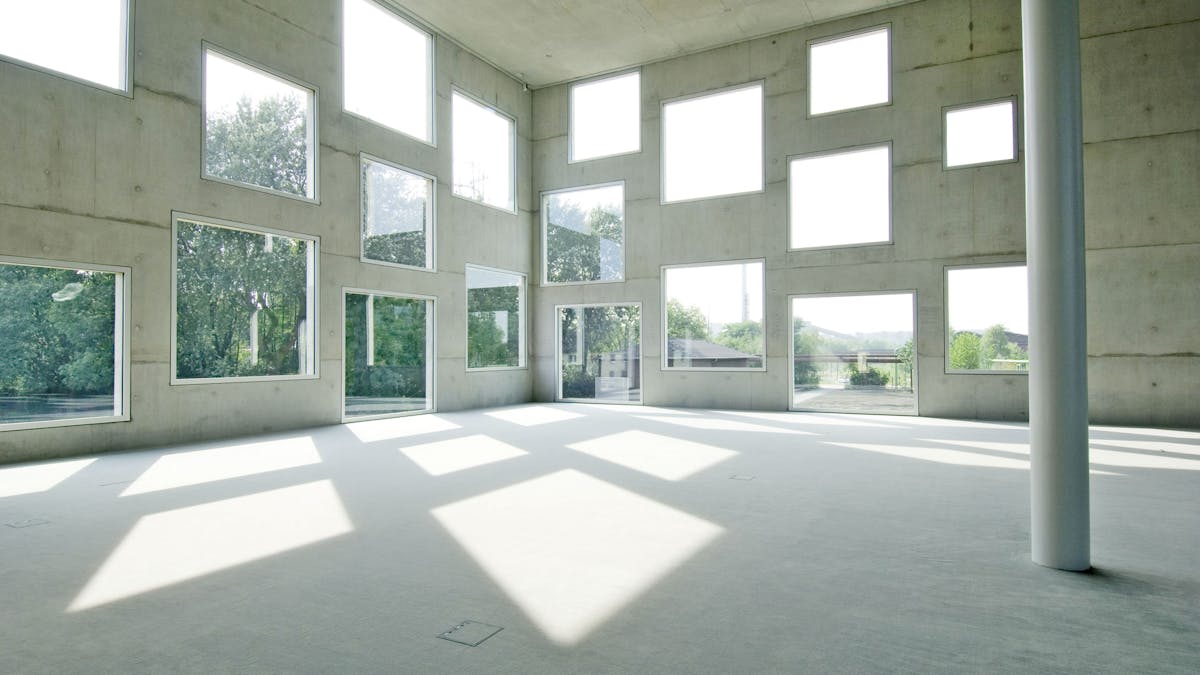 Ein großer, leerer Raum mit modernem, minimalistischem Design. Die Wände sind aus Sichtbeton, in denen zahlreiche rechteckige Fenster in unterschiedlichen Größen unregelmäßig angeordnet sind. Durch die Fenster fällt helles Tageslicht, das geometrische Schattenmuster auf dem Boden erzeugt. Eine einzelne weiße Säule steht im Raum und verleiht der Architektur eine moderne, offene Atmosphäre. Durch die Fenster sind grüne Bäume und Industrieanlagen in der Ferne zu sehen. Der Raum wirkt ruhig und lichtdurchflutet.