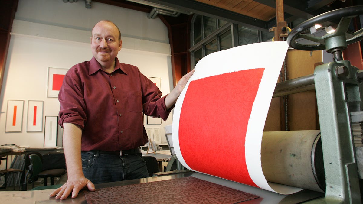 Der Künstler und Druckgrafiker Thomas Mack steht in seiner Werkstatt auf dem UNESCO-Welterbe Zollverein und präsentiert einen frisch gedruckten Linolschnitt. Er trägt ein dunkelrotes Hemd, lächelt freundlich und hebt ein großes Blatt Papier mit einem leuchtend roten Druckmotiv von einer traditionellen Druckpresse. Im Hintergrund sind an den weißen Wänden mehrere gerahmte Kunstwerke zu sehen – schlichte, geometrische Kompositionen mit roten und schwarzen Elementen, die den minimalistischen Stil seiner Werke widerspiegeln. Die Werkstatt kombiniert industrielle Ästhetik mit kreativem Schaffen: Metallrohre verlaufen unter der Decke, große Fenster lassen viel Licht in den Raum. Thomas Macks Atelier auf Zollverein ist ein Ort, an dem die handwerkliche Tradition des Drucks auf dem Gelände der ehemaligen Zeche künstlerisch weiterlebt.