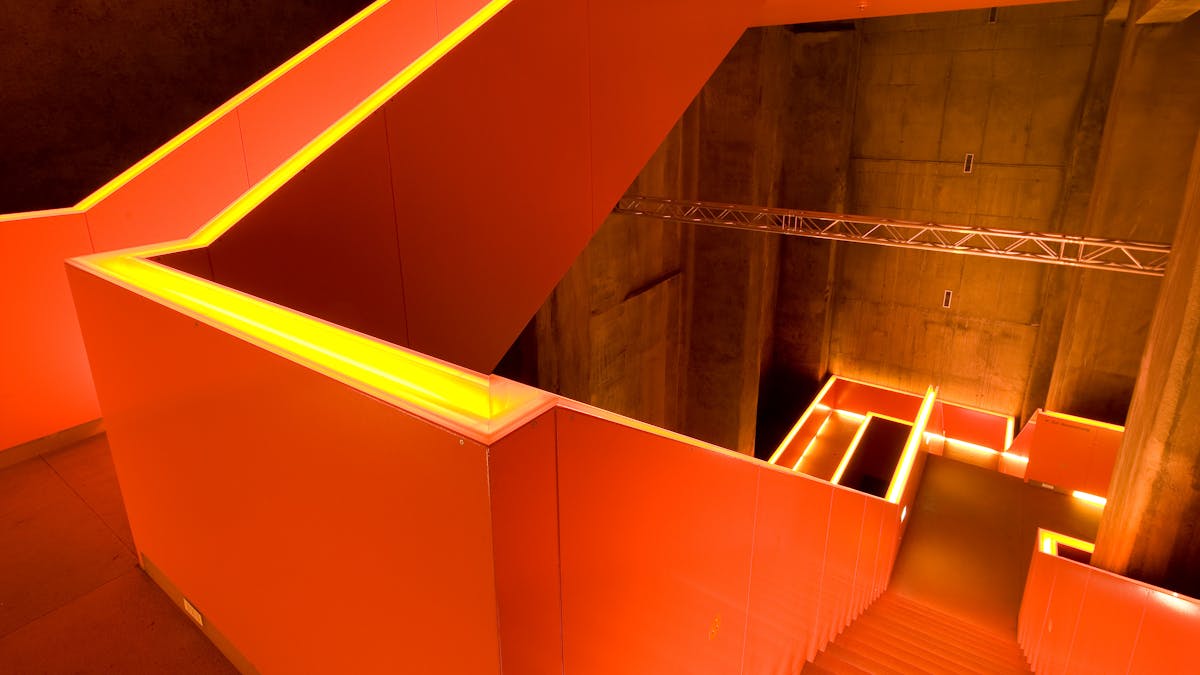 Das moderne und von Rem Koolhaas gestaltete Treppenhaus des Ruhr Museums führt in mehreren Windungen durch einen dunklen, industriellen Innenraum mit Betonwänden. Die Stufen und Geländer sind mit indirektem, orangefarbenem Licht versehen, was an einen Abstich in einem Stahlwerk erinnern soll. Die Szene vermittelt eine Mischung aus moderner Architektur und industrieller Vergangenheit.