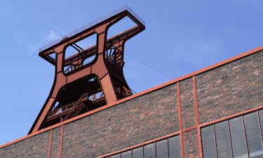 Nahaufnahme des markanten Doppelbock-Fördergerüsts der Zeche Zollverein aus einer niedrigen Perspektive. Der Blickwinkel betont die Höhe und imposante Stahlkonstruktion des ehemaligen Bergwerks. Im Vordergrund ragt eine Ziegelsteinfassade mit roten Stahlstreben und großen Fenstern empor, die den architektonischen Stil der Industrieanlage unterstreicht. Der strahlend blaue Himmel bildet einen starken Kontrast zum dunklen Backstein und der rostrot schimmernden Stahlstruktur.
