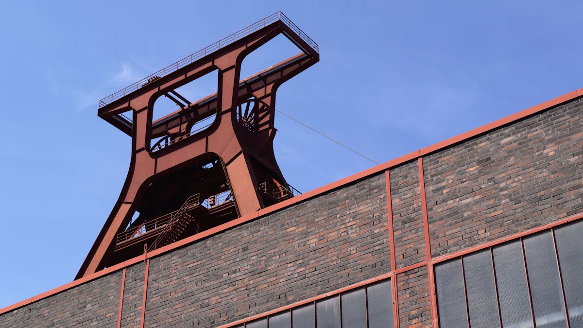 Nahaufnahme des markanten Doppelbock-Fördergerüsts der Zeche Zollverein aus einer niedrigen Perspektive. Der Blickwinkel betont die Höhe und imposante Stahlkonstruktion des ehemaligen Bergwerks. Im Vordergrund ragt eine Ziegelsteinfassade mit roten Stahlstreben und großen Fenstern empor, die den architektonischen Stil der Industrieanlage unterstreicht. Der strahlend blaue Himmel bildet einen starken Kontrast zum dunklen Backstein und der rostrot schimmernden Stahlstruktur.