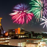 Ein beeindruckendes Feuerwerk erleuchtet den Nachthimmel über der Zeche Zollverein. Mehrere große, bunte Feuerwerkskörper explodieren in leuchtenden Farben. Im Vordergrund ist das ikonische Fördergerüst der Zeche Zollverein zu sehen, das von warmem Licht angestrahlt wird. Die umliegenden Backsteingebäude mit ihren großen Fenstern unterstützen das farbenfrohe Lichtspektakel. Unten rechts sind viele Menschen auf dem Veranstaltungsgelände zu erkennen, die das Feuerwerk genießen. Die Szene verbindet Industriekultur mit festlicher Stimmung.