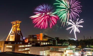 Ein beeindruckendes Feuerwerk erleuchtet den Nachthimmel über der Zeche Zollverein. Mehrere große, bunte Feuerwerkskörper explodieren in leuchtenden Farben. Im Vordergrund ist das ikonische Fördergerüst der Zeche Zollverein zu sehen, das von warmem Licht angestrahlt wird. Die umliegenden Backsteingebäude mit ihren großen Fenstern unterstützen das farbenfrohe Lichtspektakel. Unten rechts sind viele Menschen auf dem Veranstaltungsgelände zu erkennen, die das Feuerwerk genießen. Die Szene verbindet Industriekultur mit festlicher Stimmung.