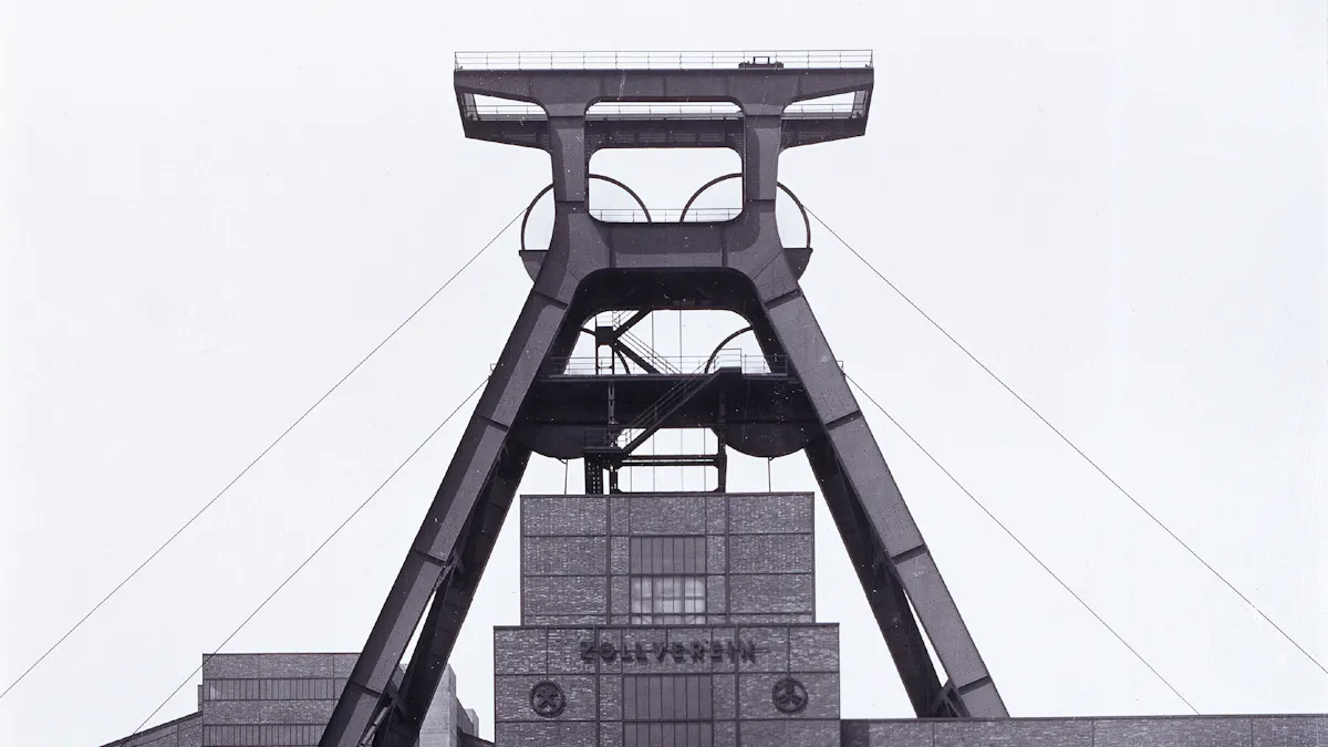 Das Bild zeigt eine ikonische Ansicht des Fördergerüsts von Schacht XII auf dem UNESCO-Welterbe Zollverein in Essen. Die Aufnahme ist in Schwarz-Weiß gehalten und hebt die klare Struktur und Architektur des Stahlgerüsts hervor. Das mächtige Doppelbock-Fördergerüst, das als eines der bekanntesten Wahrzeichen des Ruhrgebiets gilt, dominiert das Bild. Es besteht aus zwei schrägen Stahlträgern, die oben durch eine horizontale Plattform verbunden sind. Die Seile, die einst Förderkörbe in den Schacht beförderten, verlaufen schräg nach unten. Direkt unter dem Gerüst ist das Maschinenhaus mit der Aufschrift Zollverein auf der Backsteinfassade zu erkennen.