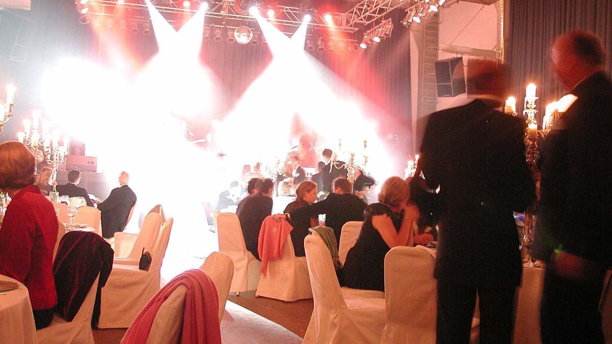 Elegante Abendveranstaltung mit festlich gedeckten Tischen und weiß verhüllten Stühlen. Gäste in Abendgarderobe sitzen und unterhalten sich, während im Hintergrund eine Bühne mit heller Scheinwerferbeleuchtung erstrahlt. Die Atmosphäre wird durch große, goldene Kerzenleuchter verstärkt, die auf den Tischen stehen. Die Beleuchtung ist warm, und die Lichtstrahlen der Bühnenscheinwerfer durchziehen den Raum. Eine lebendige und festliche Szene mit einem luxuriösen Ambiente.