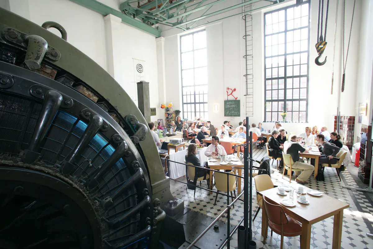 Das Bild zeigt das Café Zollverein, das sich in der ehemaligen Maschinenhalle der Schachtanlage 3/7/10 auf dem UNESCO-Welterbe Zollverein in Essen befindet. Das Café kombiniert industrielle Architektur mit einer gemütlichen Gastronomie-Atmosphäre. Der Raum ist geprägt von hohen weißen Wänden, großen Fenstern mit schwarzem Stahlrahmen, gewaltigen Maschinen, einem schwarz-weiß gefliesten Boden und grünen Stahlträgern an der Decke. Von der Decke hängen alte Industriekräne mit schweren Eisenhaken, die an die frühere Nutzung der Halle erinnern. Im Raum verteilt stehen Holztische mit verschiedenen Stühlen, an denen Gäste essen, trinken und sich unterhalten. Das Café ist gut besucht, Menschen in unterschiedlichen Altersgruppen genießen ihre Mahlzeiten. Im Hintergrund gibt es eine kleine Theke mit Dekorationen sowie eine Tafel mit Kreideschrift, auf der Willkommen im Café Zollverein steht. Ein rotes Logo mit einer tanzenden Figur verweist auf das angrenzende Phänomania Erfahrungsfeld, ein Mitmachmuseum für Naturwissenschaften.