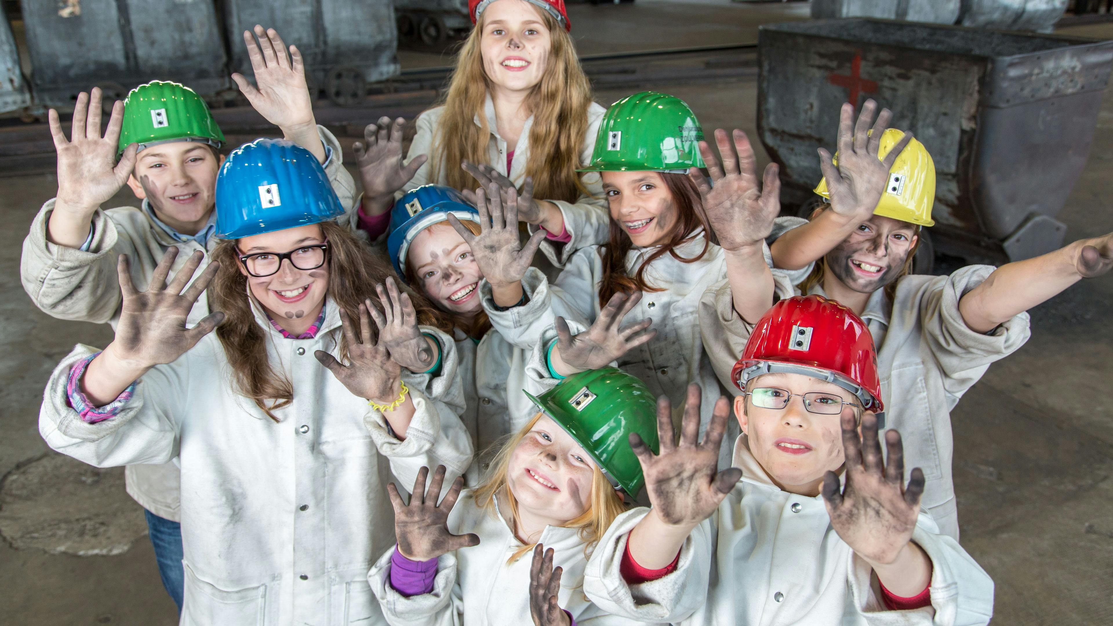 Eine fröhliche Gruppe von Kindern in Bergmannsjacken und bunten Schutzhelmen zeigt lachend ihre mit Kohlenstaub verschmutzten Hände in die Kamera. Die Kinder nehmen an einem interaktiven Erlebnisprogramm auf dem Gelände der Zeche Zollverein teil, bei dem sie spielerisch in die Welt des Bergbaus eintauchen. Im Hintergrund sind alte Förderwagen zu sehen, die an die historische Nutzung des Areals erinnern. Die Szene vermittelt Begeisterung und Entdeckerfreude, während die Kinder die Geschichte und Arbeit der Bergleute hautnah erleben.