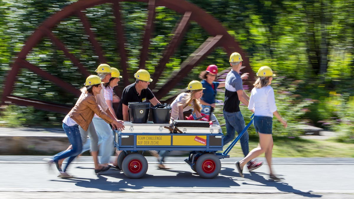 Das Bild zeigt eine Gruppe von Teilnehmern mit gelben Schutzhelmen, die während einer Teambuilding-Veranstaltung auf dem Gelände des UNESCO-Welterbes Zollverein einen Handkarren mit Kisten und Werkzeugen ziehen und schieben. Die Veranstaltung trägt den Titel Team Challenge auf der Zeche, wie auf einem Schild am Karren zu lesen ist. Die Gruppe bewegt sich dynamisch durch den Zollverein Park, der durch grüne Bäume und eine große historische Förderrad-Skulptur im Hintergrund geprägt ist. Die Teilnehmenden sind engagiert und in Bewegung, was den sportlichen und kooperativen Charakter der Aktivität unterstreicht. Diese Art von Veranstaltungen verbindet spielerische Herausforderungen mit der industriellen Geschichte Zollvereins, um Teamarbeit und Zusammenhalt zu stärken.
