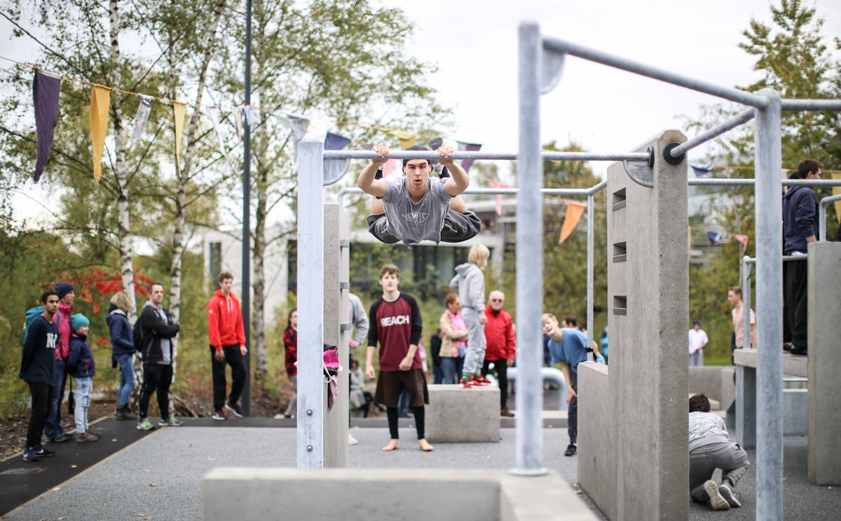 Ein junger Sportler führt eine akrobatische Übung an einer horizontalen Stange im Zollverein Park aus. Er hält sich mit den Händen an der Stange fest und hebt seinen Körper parallel zum Boden, während seine Beine gespreizt in der Luft schweben. Um ihn herum beobachten mehrere Menschen das Geschehen, einige tragen sportliche Kleidung, andere sind als Zuschauer gekommen. Der Outdoor-Sportbereich ist mit Betonblöcken, Stangen und Kletterelementen ausgestattet, ideal für Parkour und Calisthenics. Im Hintergrund sind grüne Bäume, bunte Wimpelketten und moderne Gebäude zu sehen. Die Szene vermittelt eine dynamische Atmosphäre inmitten der Industriekulisse von Zollverein.