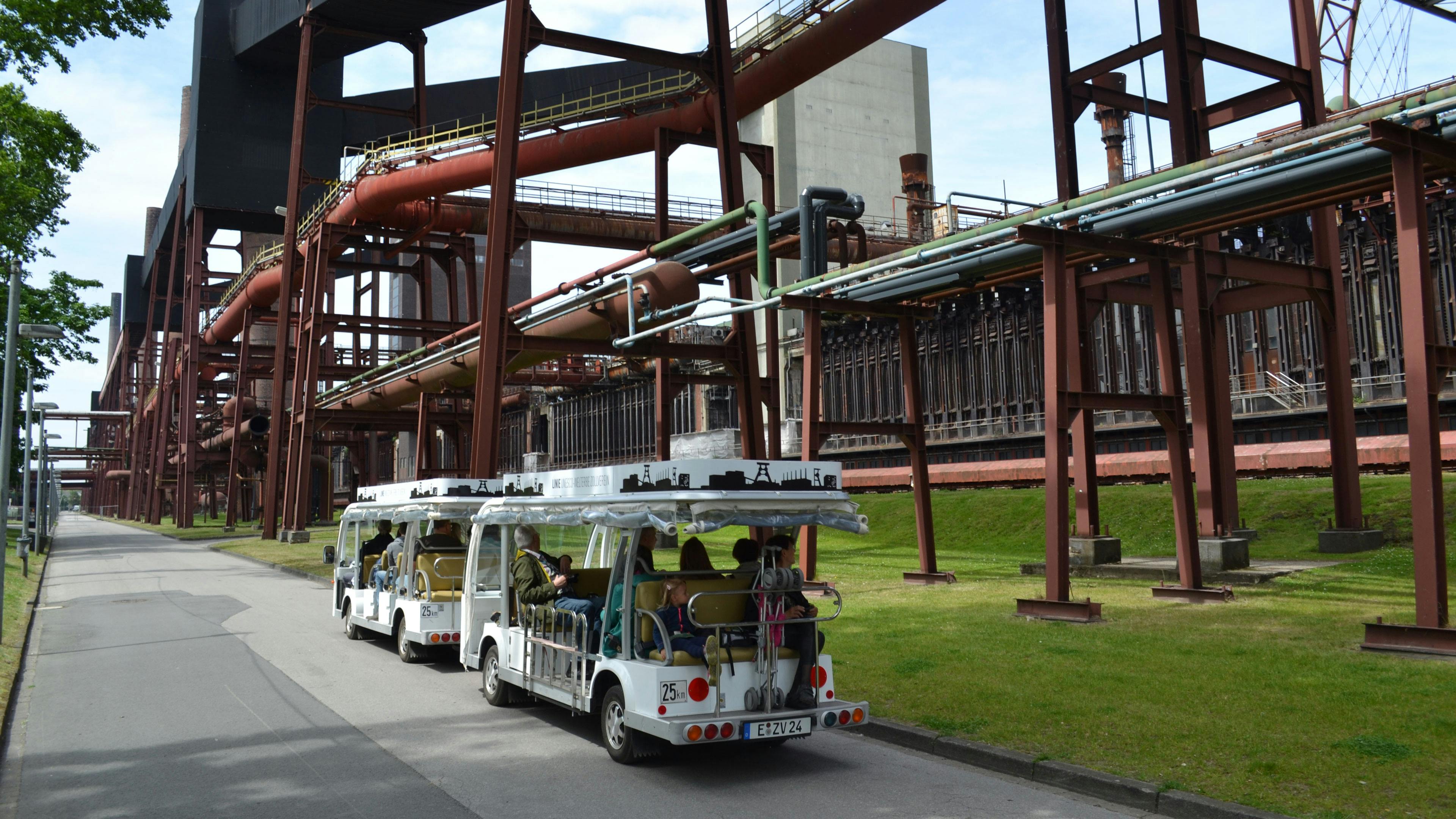 Das Bild zeigt einen offenen Elektrobus der Zollverein-Rundfahrten, der Besucher durch das weitläufige Gelände des UNESCO-Welterbes Zollverein fährt. Der Bus ist weiß, hat eine offene Bauweise mit einem transparenten Dach und zeigt auf dem Dach eine Silhouette der Fördergerüste und Industrieanlagen des Ruhrgebiets. Im Hintergrund sind die rostroten Stahlkonstruktionen und Rohrleitungen der Kokerei Zollverein zu sehen, die einst ein zentraler Ort der Kohleverarbeitung war. Die Besucher im Bus sitzen entspannt und genießen die Fahrt, während sie sich von einem Gästeführer die Geschichte und Bedeutung der Industrieanlage erklären lassen. Diese barrierefreie Rundfahrt ist besonders für Menschen mit eingeschränkter Mobilität geeignet und ermöglicht von Frühjahr bis Herbst einen umfassenden Eindruck der ehemaligen Zeche und Kokerei.