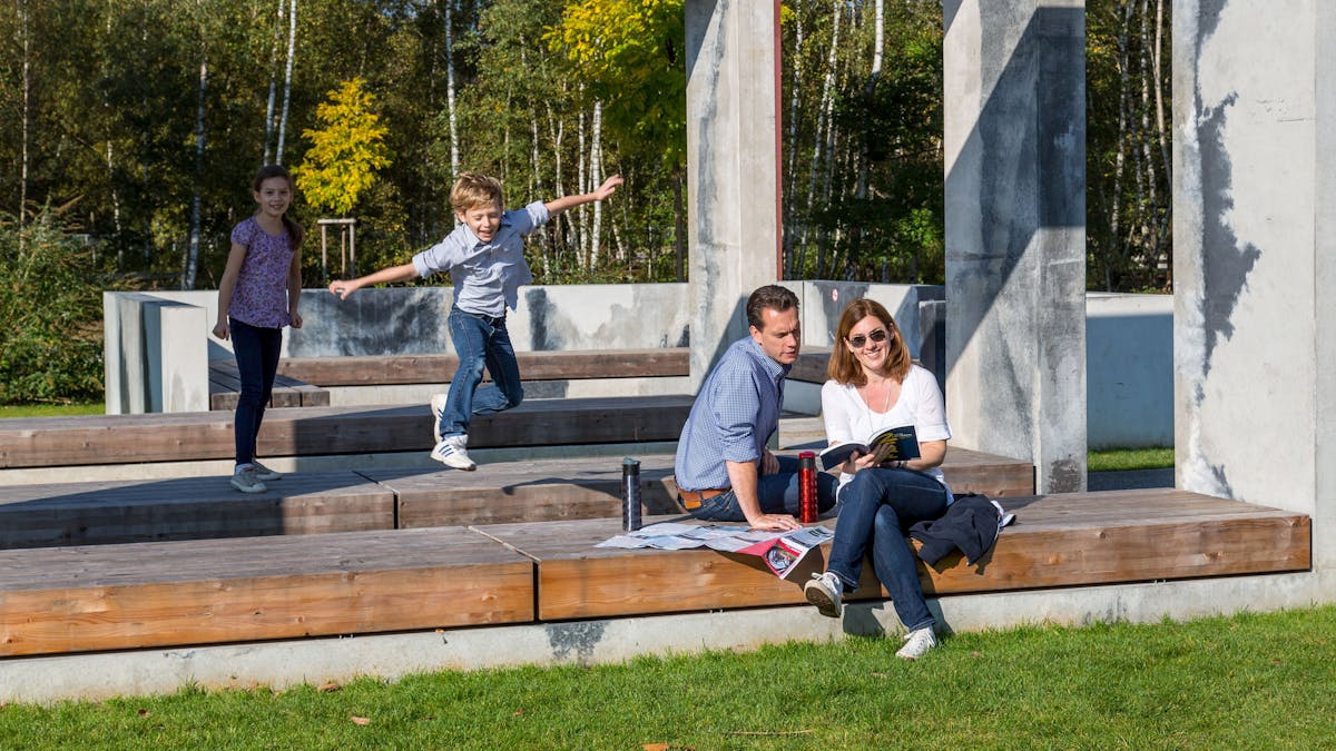 Eine Familie verbringt einen sonnigen Tag im Zollverein Park. Im Vordergrund sitzt ein Paar entspannt auf einer hölzernen Sitzbank, umgeben von modernen Betonelementen. Die Frau liest ein Buch, während der Mann interessiert auf eine ausgebreitete Landkarte schaut. Zwei Kinder spielen fröhlich auf den Bänken im Hintergrund: Ein Junge springt mit ausgebreiteten Armen durch die Luft, während ein Mädchen ihn lachend beobachtet. Der Park ist von grünen Wiesen, Bäumen und einer naturnahen Umgebung umgeben, die das ehemalige Industriegelände in einen Erholungsort verwandelt.