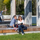 Eine Familie verbringt einen sonnigen Tag im Zollverein Park. Im Vordergrund sitzt ein Paar entspannt auf einer hölzernen Sitzbank, umgeben von modernen Betonelementen. Die Frau liest ein Buch, während der Mann interessiert auf eine ausgebreitete Landkarte schaut. Zwei Kinder spielen fröhlich auf den Bänken im Hintergrund: Ein Junge springt mit ausgebreiteten Armen durch die Luft, während ein Mädchen ihn lachend beobachtet. Der Park ist von grünen Wiesen, Bäumen und einer naturnahen Umgebung umgeben, die das ehemalige Industriegelände in einen Erholungsort verwandelt.