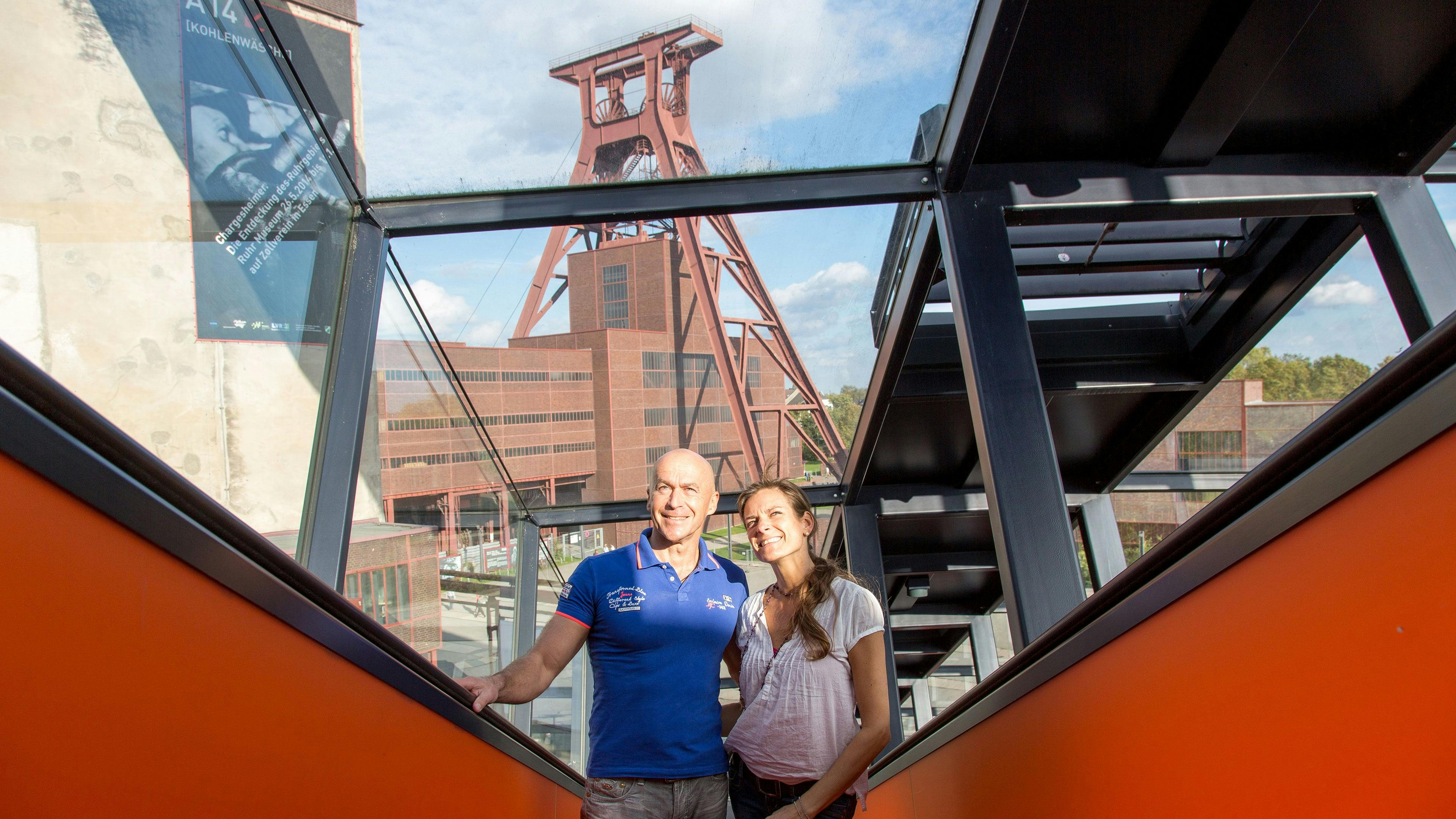 Ein lächelndes Paar steht auf der markanten orangefarbenen Rolltreppe der Kohlenwäsche auf dem Gelände der Zeche Zollverein in Essen. Die Rolltreppe, die als längste freistehende Rolltreppe Deutschlands gilt, führt hinauf zum Ruhr Museum und ist von schwarzem Stahl und Glas eingefasst. Durch die verglasten Seiten fällt der Blick auf das ikonische Fördergerüst von Schacht XII, das sich imposant vor blauem Himmel erhebt. Das Paar genießt die Aussicht auf die denkmalgeschützte Industriekulisse des Weltkulturerbes Zollverein.