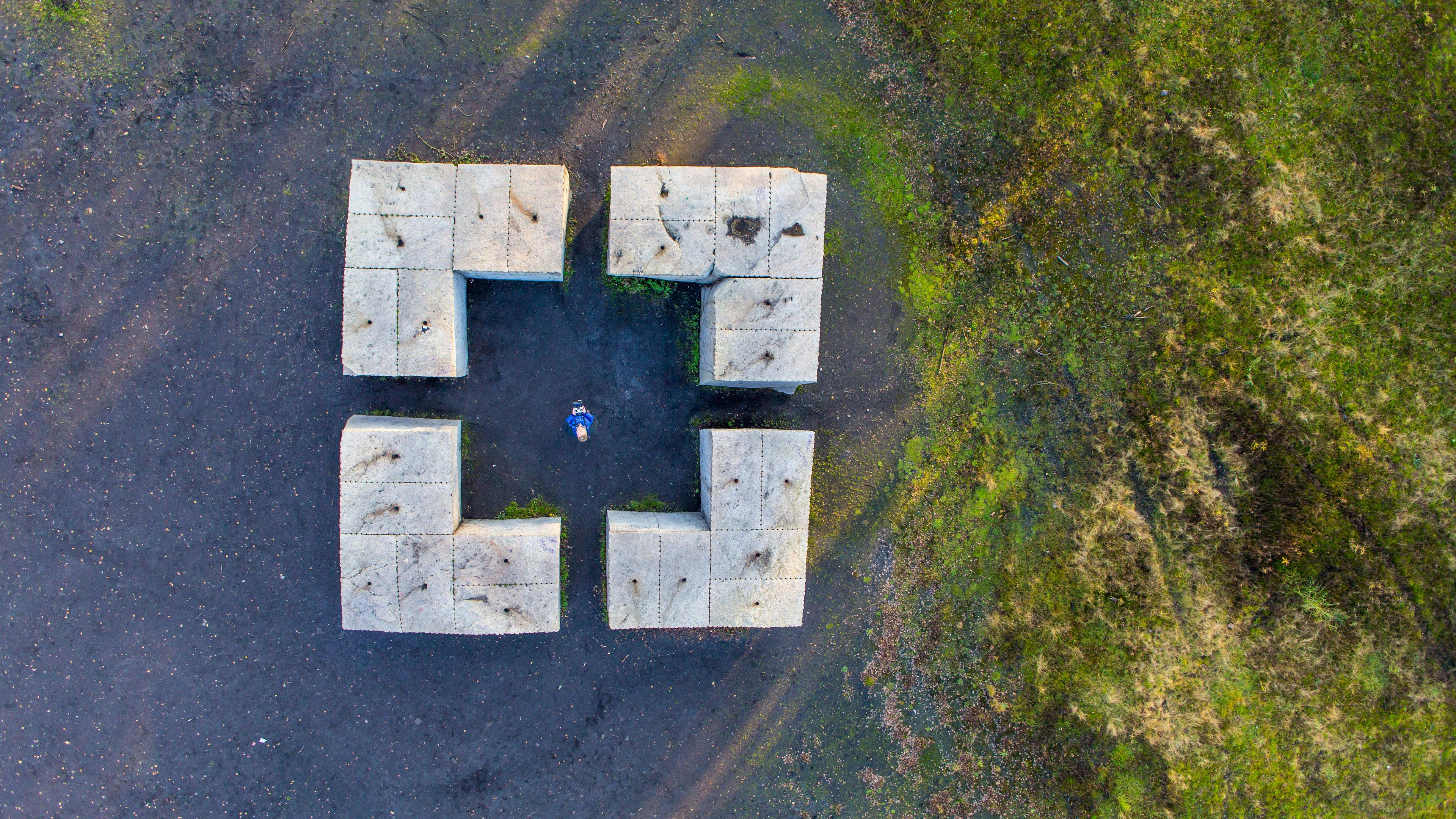 Luftaufnahme der Skulptur Castell von Ulrich Rückriem im Zollverein Park. Die monumentale Steinanlage besteht aus vier symmetrisch angeordneten L-förmigen Granitblöcken, die eine offene quadratische Struktur bilden. Die Skulptur steht auf dunklem Boden, umgeben von grüner Natur. In der Mitte der Anordnung steht eine Person in blauer Kleidung, die nach oben schaut, was die imposante Größe der Steine betont. Das Werk ist Teil der künstlerischen Umgestaltung des ehemaligen Industrieareals und lädt Besucher dazu ein, es zu betreten und aus verschiedenen Perspektiven zu erleben.
