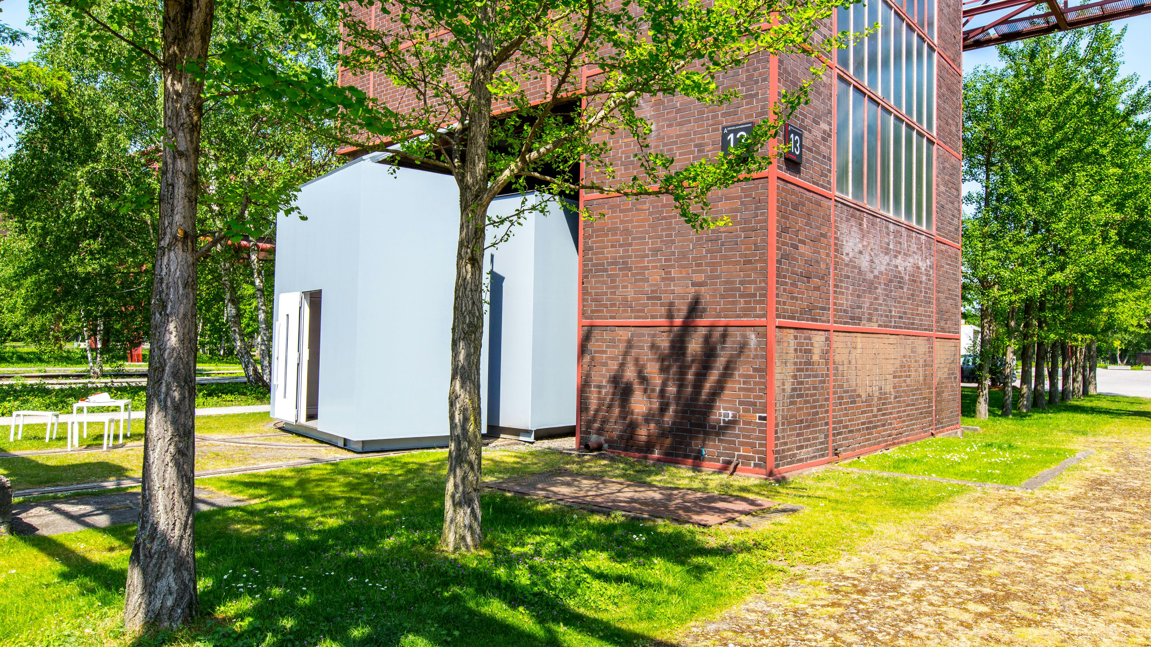Der Eingangsbereich zum Kesselaschebunker auf dem Gelände der Zeche Zollverein. Vor dem historischen Backsteinbau mit großen Fensterflächen steht eine moderne, weiße Eingangskonstruktion. Die Umgebung ist von grünen Rasenflächen und Bäumen geprägt, die Schatten auf den Boden werfen. Im Vordergrund befinden sich einige weiße Sitzgelegenheiten. Der Kesselaschebunker war einst ein Teil der Industrieanlage und wird heute für verschiedene Zwecke genutzt.