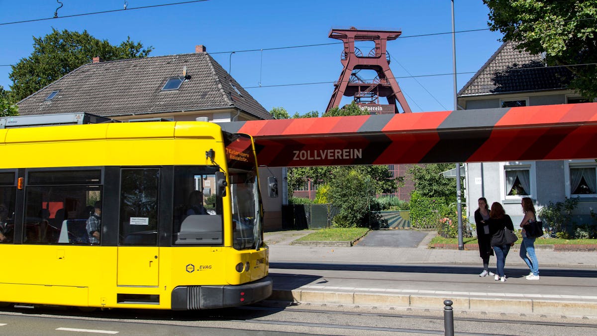 Eine gelbe Straßenbahn der Linie 107 hält an der Haltestelle Zollverein in Essen. Die Haltestelle ist mit einer markanten, rot-schwarz gestreiften Überdachung versehen, auf der in großen weißen Buchstaben Zollverein steht. Im Hintergrund ragt das berühmte Fördergerüst der Zeche Zollverein über die umliegenden Häuser hinaus. Drei Frauen stehen auf dem Bahnsteig und unterhalten sich. Die Szene zeigt die Verbindung zwischen dem UNESCO-Welterbe Zollverein und dem öffentlichen Nahverkehr der Stadt Essen.