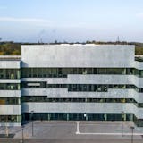 Die moderne Fassade des Neubaus der Folkwang Universität der Künste auf dem Gelände der Zeche Zollverein in Essen. Das Gebäude zeichnet sich durch seine klare, geometrische Architektur aus, mit einer hellen Steinverkleidung und großen Fensterfronten. Vor dem Haupteingang sind drei orangefarbene Fahnen mit dem Schriftzug Folkwang aufgestellt. Das Gebäude befindet sich inmitten der historischen Industrieanlage und steht für die Verbindung von Kunst, Design und industriekulturellem Erbe auf Zollverein.