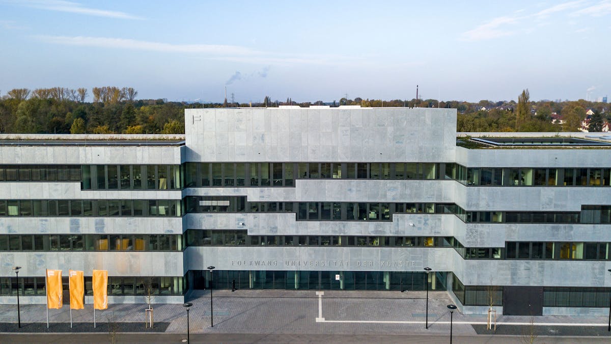 Die moderne Fassade des Neubaus der Folkwang Universität der Künste auf dem Gelände der Zeche Zollverein in Essen. Das Gebäude zeichnet sich durch seine klare, geometrische Architektur aus, mit einer hellen Steinverkleidung und großen Fensterfronten. Vor dem Haupteingang sind drei orangefarbene Fahnen mit dem Schriftzug Folkwang aufgestellt. Das Gebäude befindet sich inmitten der historischen Industrieanlage und steht für die Verbindung von Kunst, Design und industriekulturellem Erbe auf Zollverein.