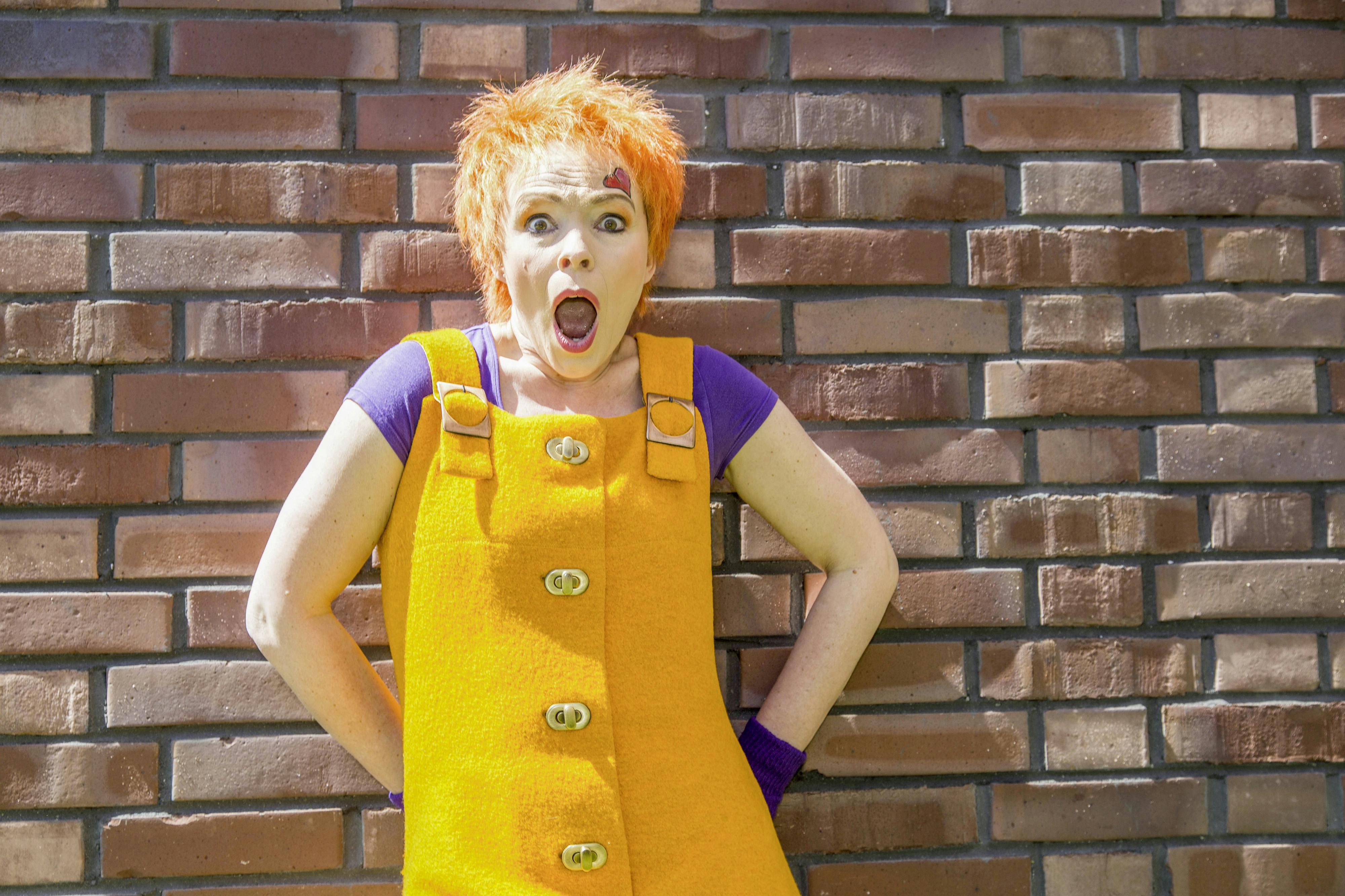 Das Bild zeigt eine farbenfrohe Figur vor einer roten Backsteinmauer, die als Hexe Kleinlaut bekannt ist. Sie trägt ein auffälliges gelbes Kleid mit großen Holzknöpfen und lila Shirt darunter. Ihre kurzen, leuchtend orangefarbenen Haare unterstreichen ihren verspielten, energiegeladenen Ausdruck. Ihr Gesicht zeigt eine überraschte oder begeisterte Mimik mit weit aufgerissenen Augen und offenem Mund. Auf ihrer Stirn ist ein kleines rotes Herz aufgemalt. Hexe Kleinlaut ist eine bekannte Kinderfigur, die auf dem Gelände von Zollverein bei Veranstaltungen oder kulturellen Programmen für junge Besucher auftritt. Ihr auffälliges, fröhliches Erscheinungsbild und ihre ausdrucksstarke Körpersprache sorgen für eine lebendige, interaktive Unterhaltung, die perfekt in das kreative und kulturelle Konzept von Zollverein passt.