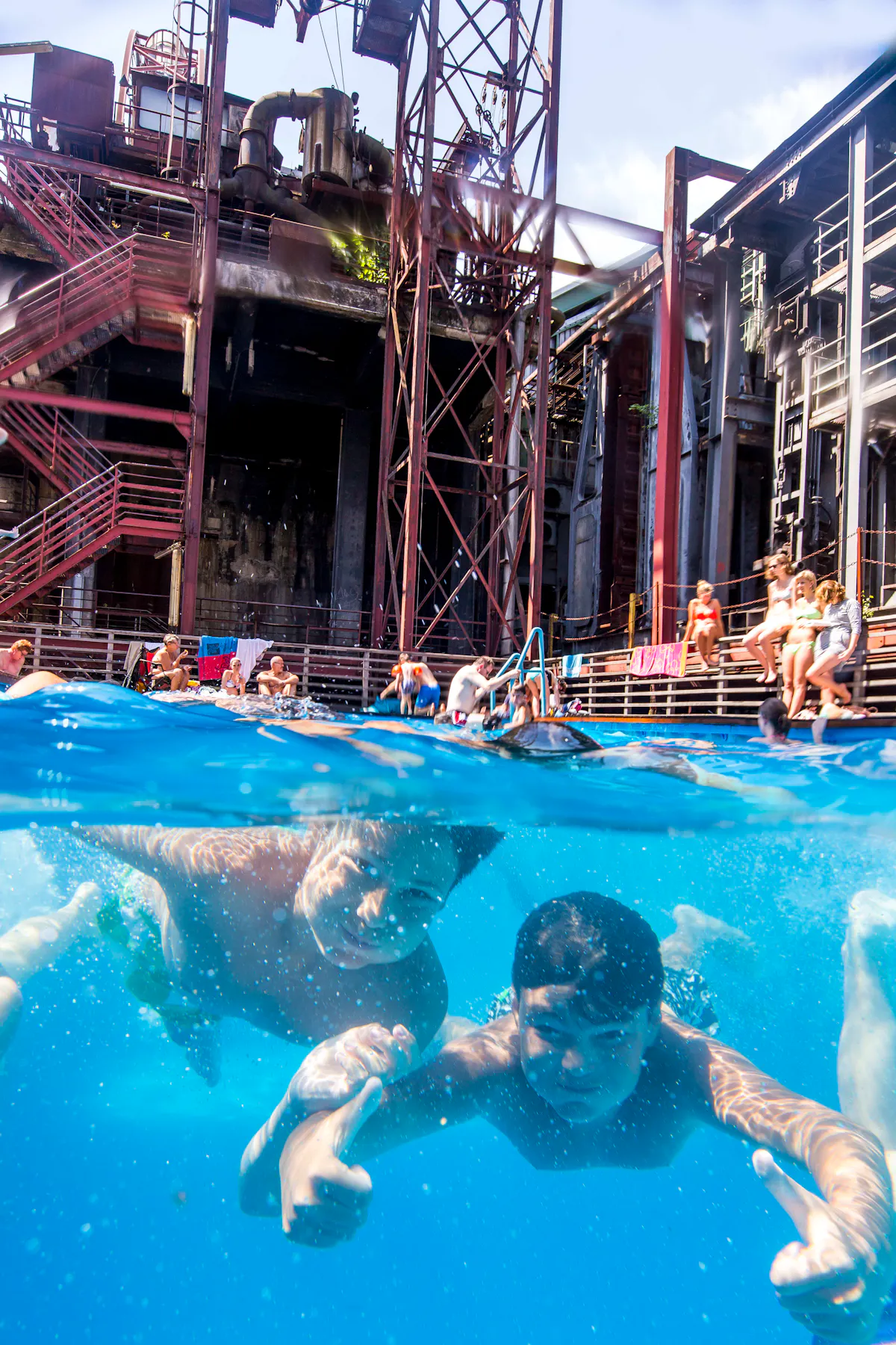 Das Bild zeigt eine sommerliche Szene im Werksschwimmbad auf der Kokerei Zollverein, einem interaktiven Kunstprojekt, das während der Sommerferien inmitten der industriellen Kulisse der ehemaligen Kokerei geöffnet ist. Das Foto ist in einem besonderen Perspektivstil aufgenommen – es ist zur Hälfte unter Wasser und zur Hälfte über Wasser. Unterhalb der Wasserlinie schwimmen zwei junge Badegäste auf die Kamera zu und zeigen lachend Daumen hoch. Das Wasser ist klar und reflektiert die Sonne, wodurch ein lebendiges, erfrischendes Bild entsteht. Über der Wasserlinie ist die industrielle Umgebung der Kokerei sichtbar: rostige Stahlgerüste, alte Rohre und rote Treppenanlagen bilden einen starken Kontrast zur Freizeitaktivität im Becken. Auf den Tribünen rund um das Becken sitzen und liegen zahlreiche Badegäste in Badekleidung, einige trocknen sich mit Handtüchern ab, andere unterhalten sich oder beobachten das Treiben im Wasser.